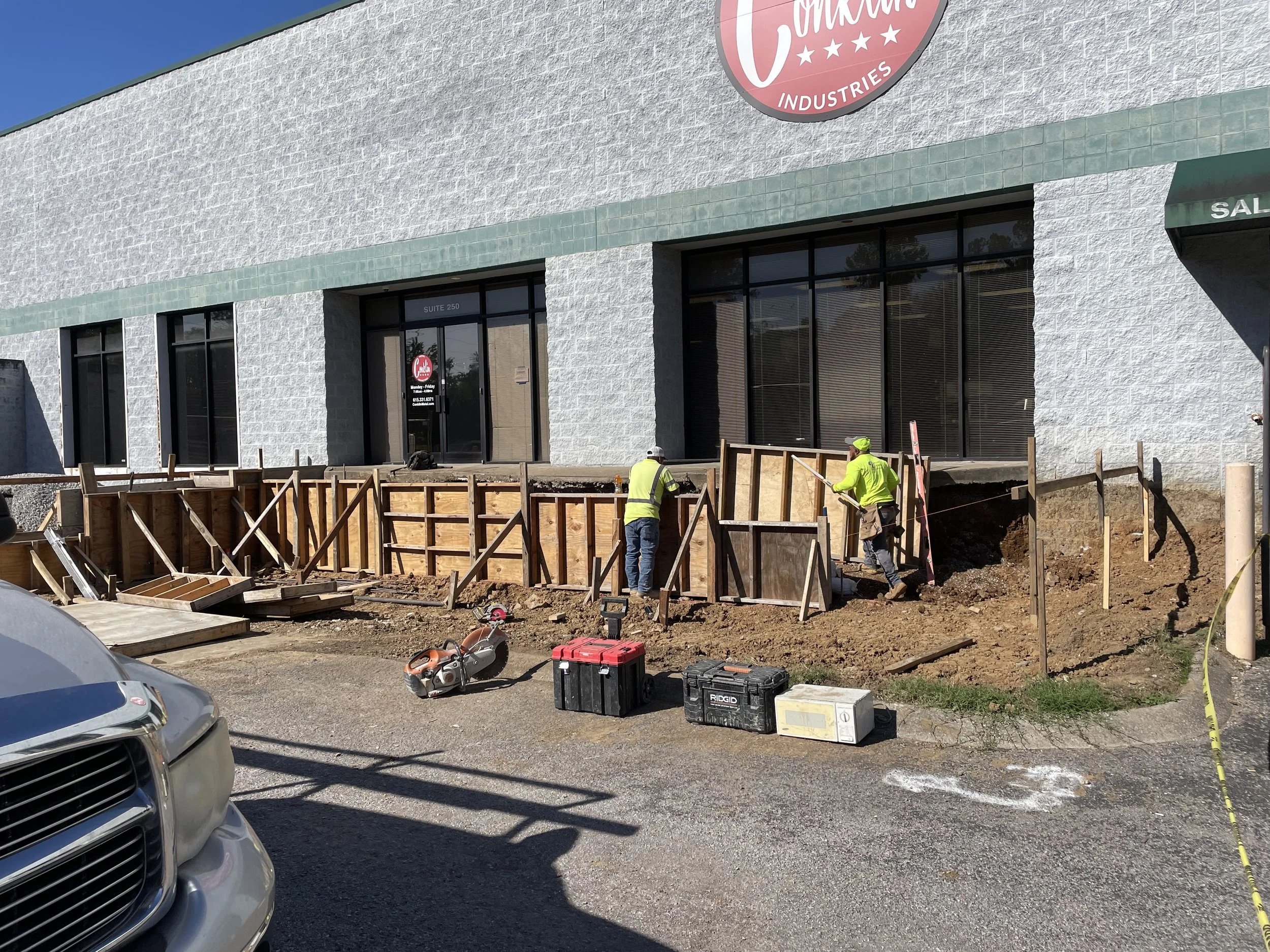 Construction workers in yellow safety vests working on sidewalk renovation outside a commercial building with large windows and a sign that reads "Cimmco Industries". Tools and equipment are on the ground.
