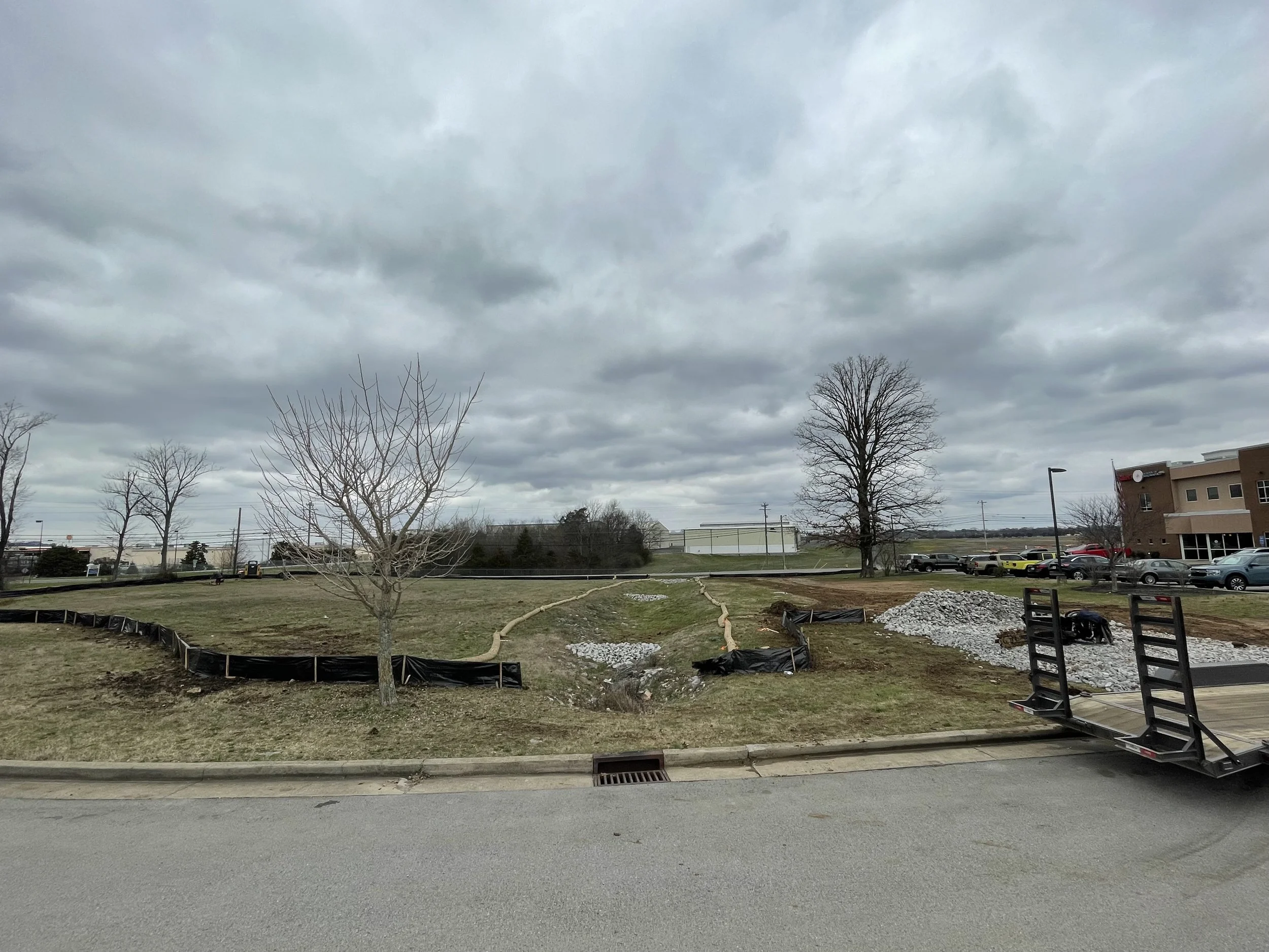 A mostly cloudy sky over a patch of grass with newly planted trees and construction materials, including black fabric and gravel, near a parking lot with cars and a building in the background.