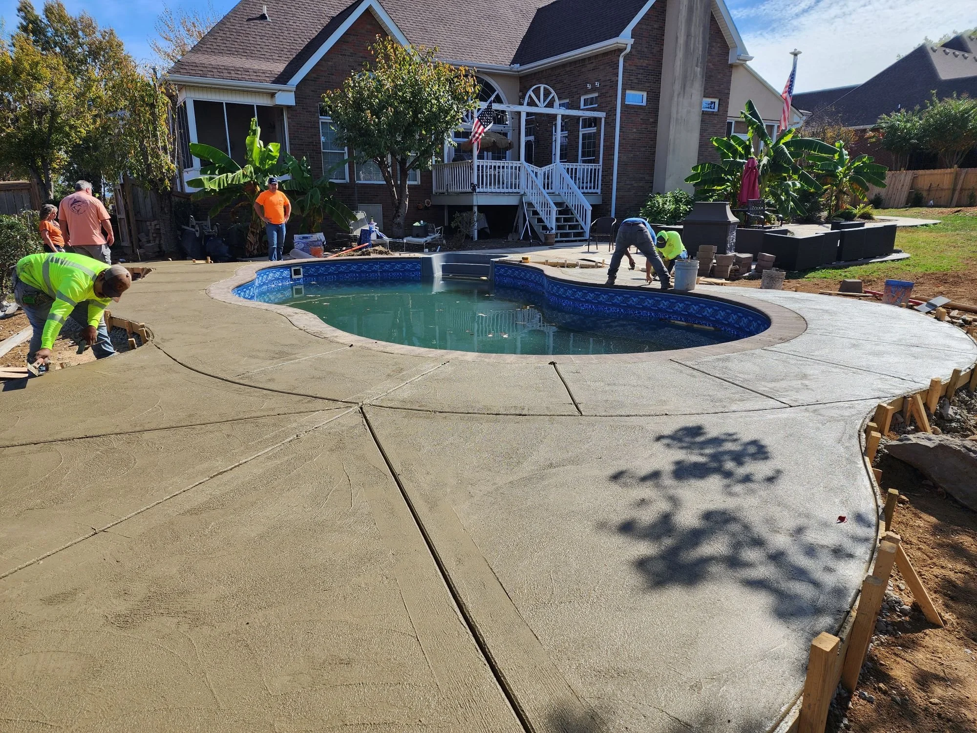 Construction workers are pouring and smoothing concrete around a backyard swimming pool, with a house and patio in the background.