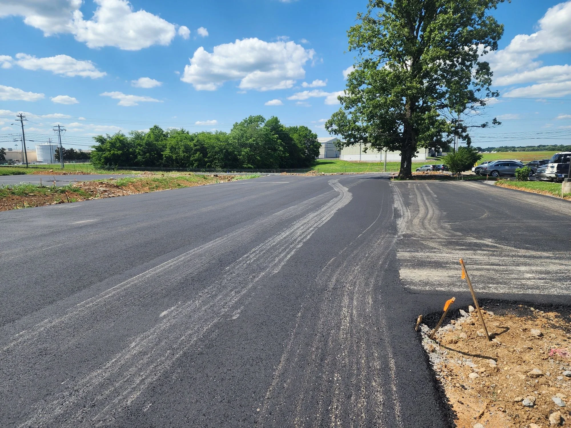 Newly paved asphalt parking lot with a large tree and a partly cloudy sky.
