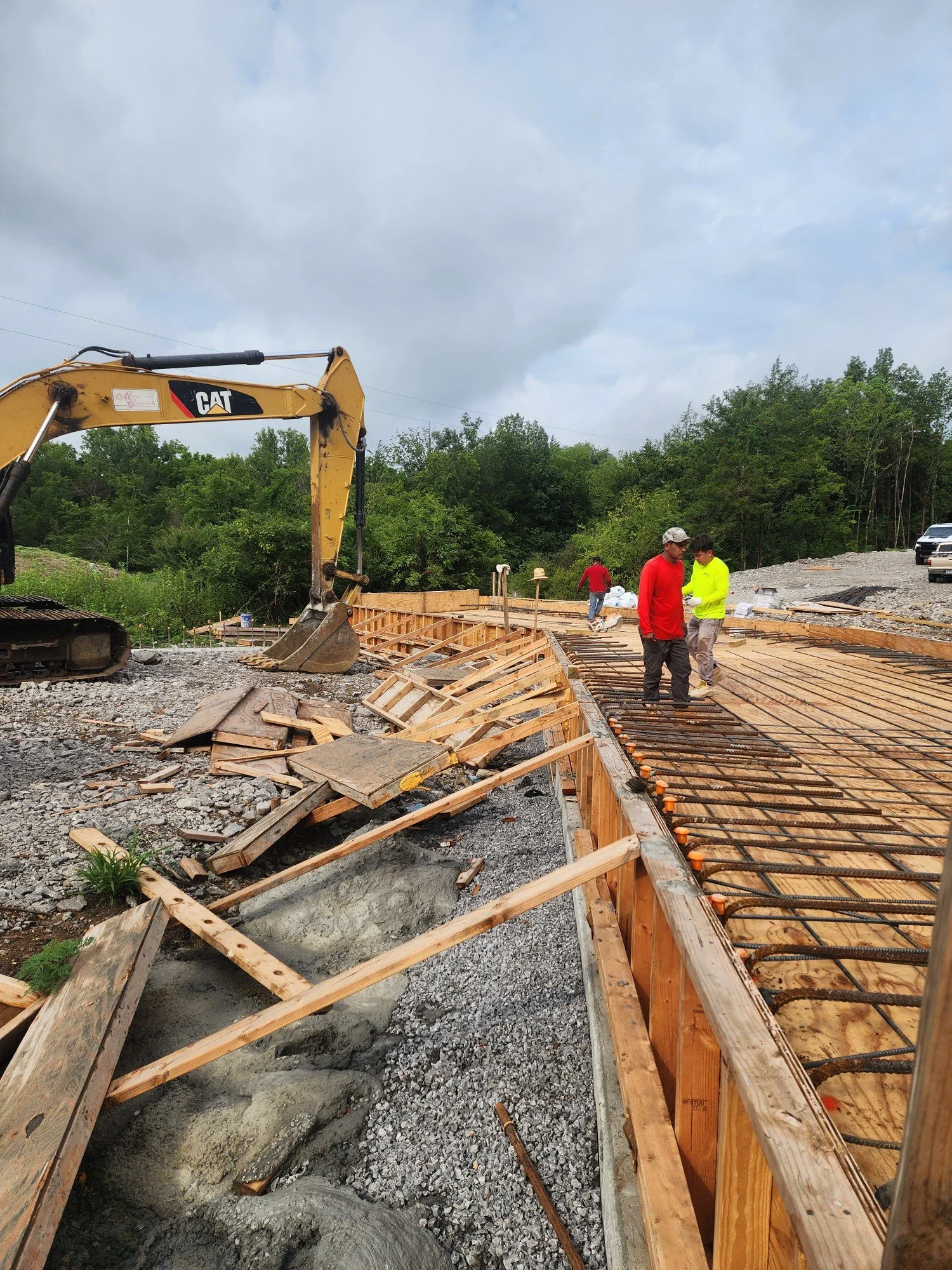 Construction site with wooden framework, rebar, and an excavator, workers in safety gear, and a cloudy sky.