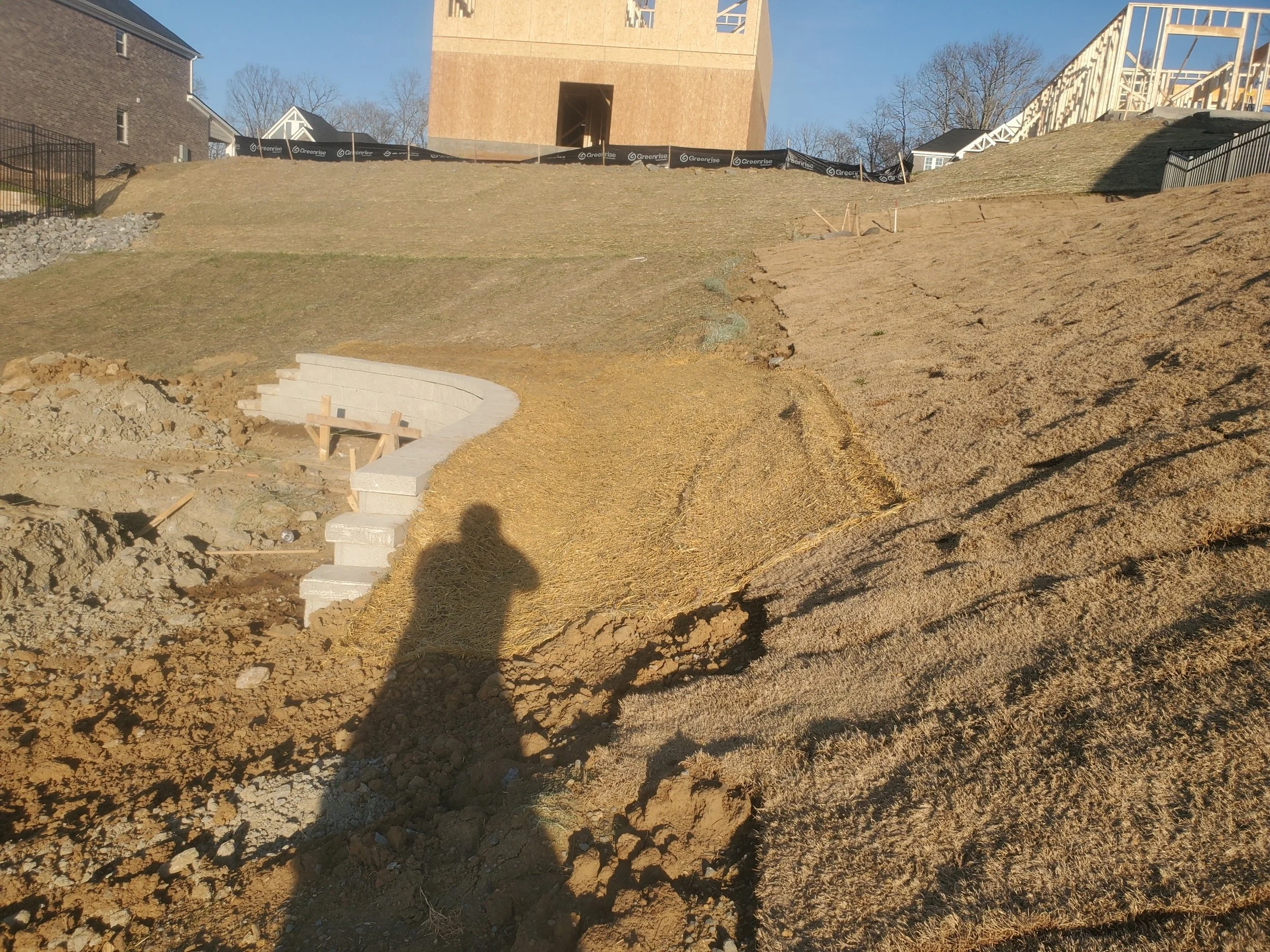 Construction site on a hill with concrete steps and wooden framing, with partially disturbed soil and grass, and residential houses in the background.
