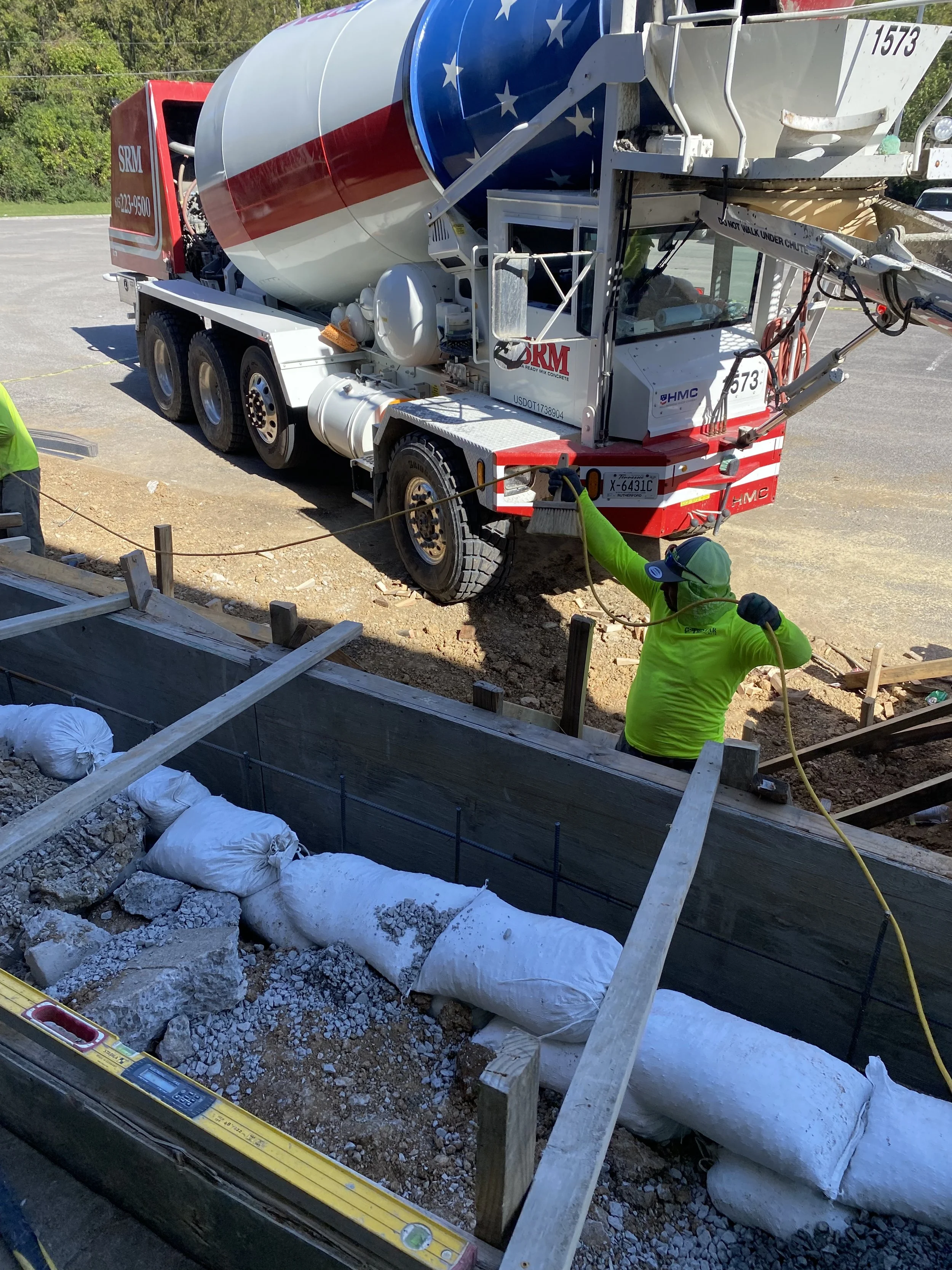 A construction worker in a bright yellow-green shirt and green cap working at a construction site with a concrete mixer truck parked nearby.