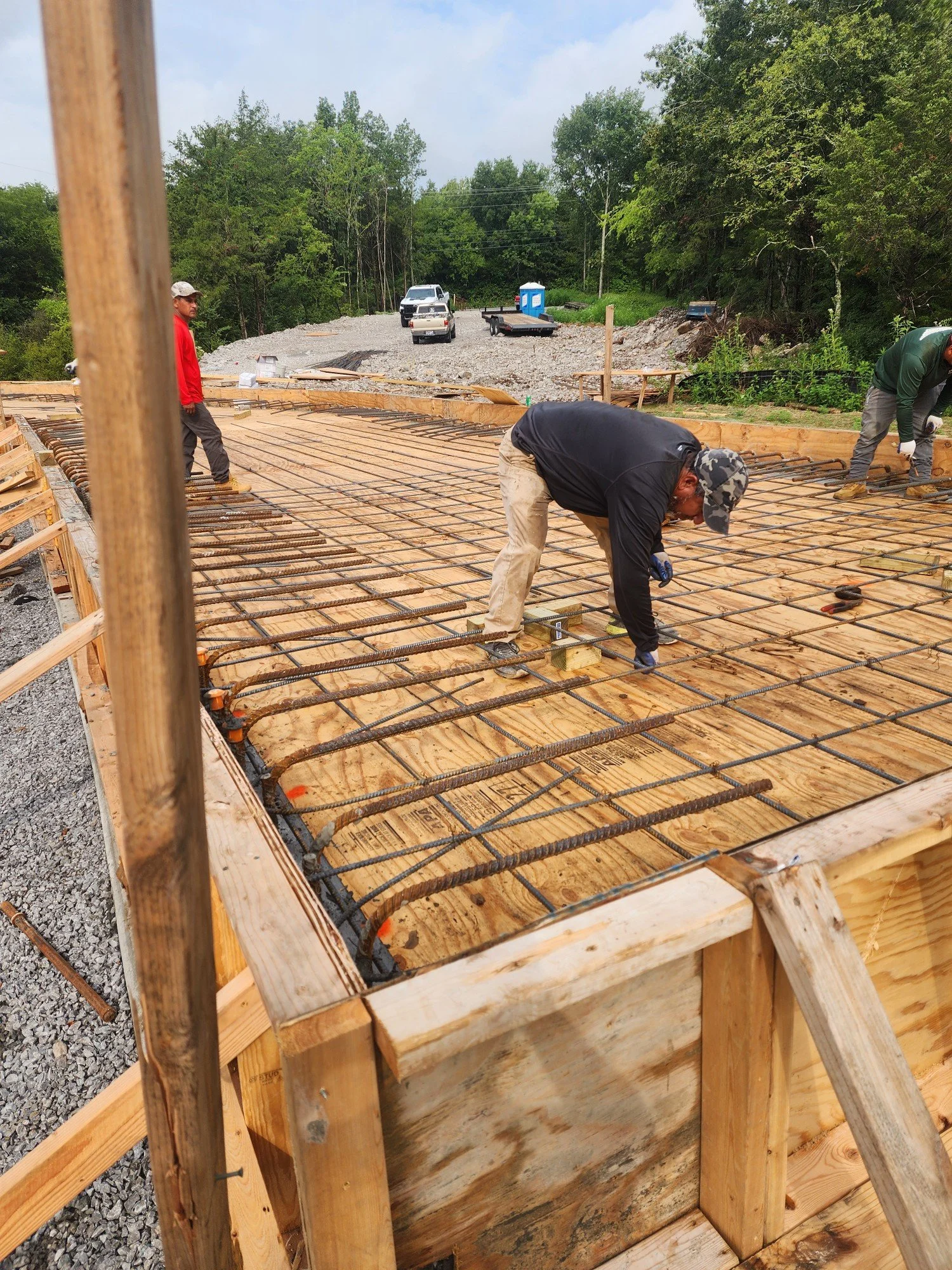 Construction workers are working on a building foundation with rebar reinforcement, surrounded by wooden formwork, on a partly cloudy day in a wooded area.