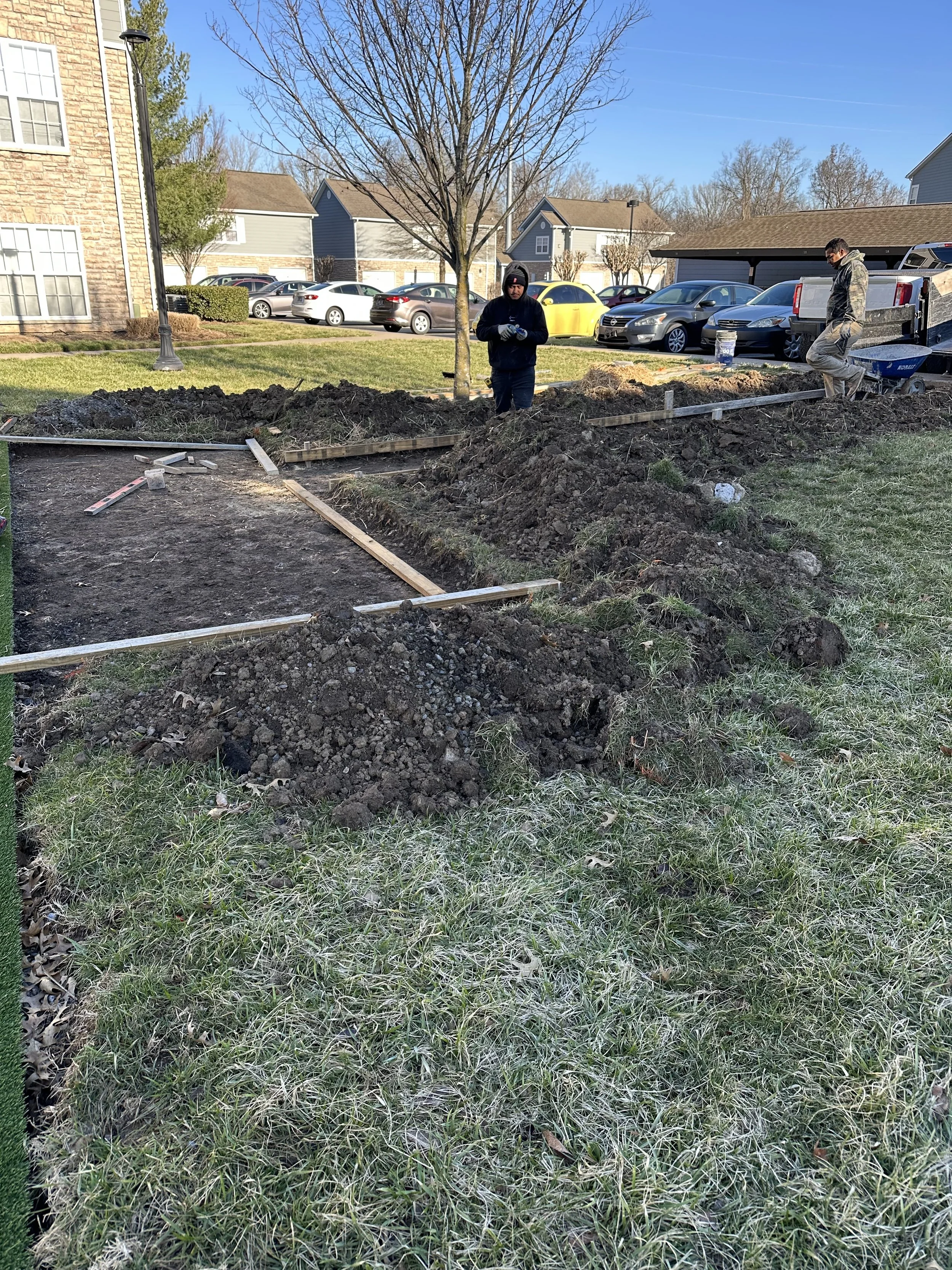 People working on landscaping or construction project in a grassy area with dirt, rocks, and wooden frames, in a residential neighborhood with cars parked in the background.