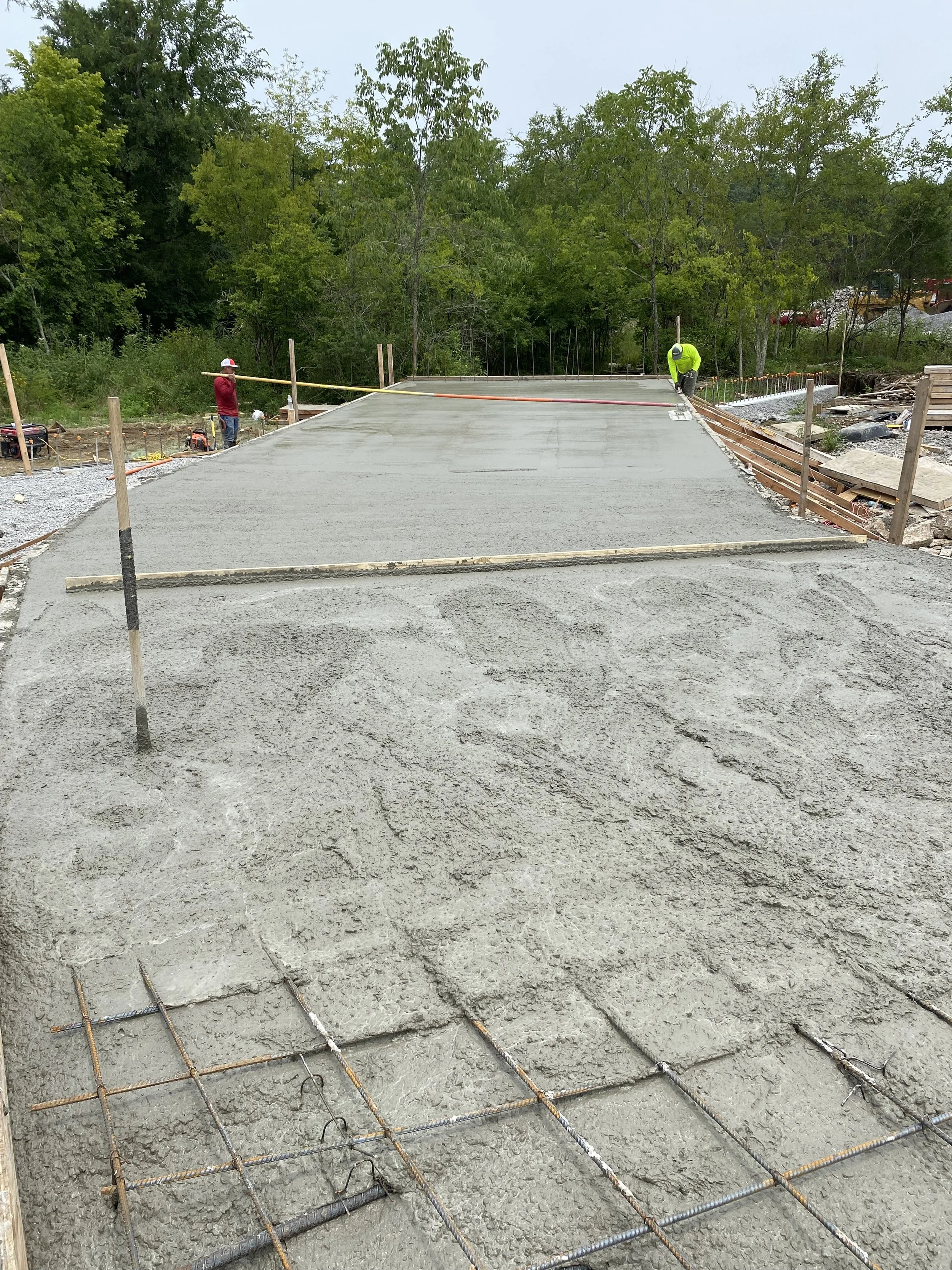 Construction workers pouring and smoothing concrete on a building site outdoors, with a partially finished concrete slab, green trees in the background, and construction tools and materials around.
