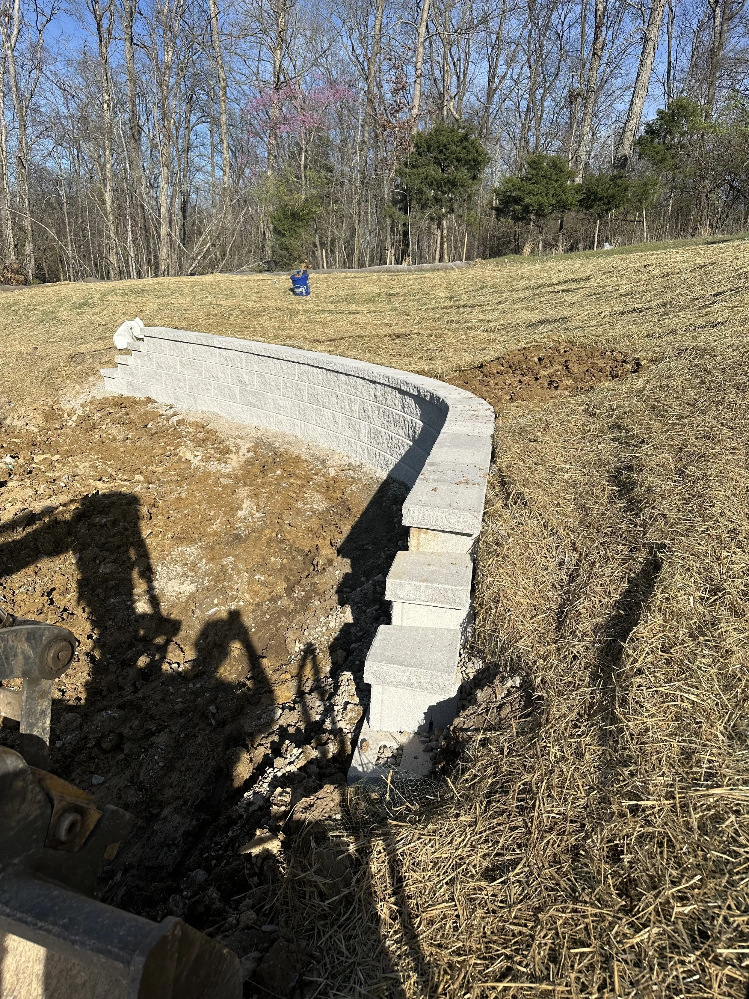 Construction site with a new gray brick retaining wall being built on a grassy hillside, with a background of leafless trees and clear blue sky.