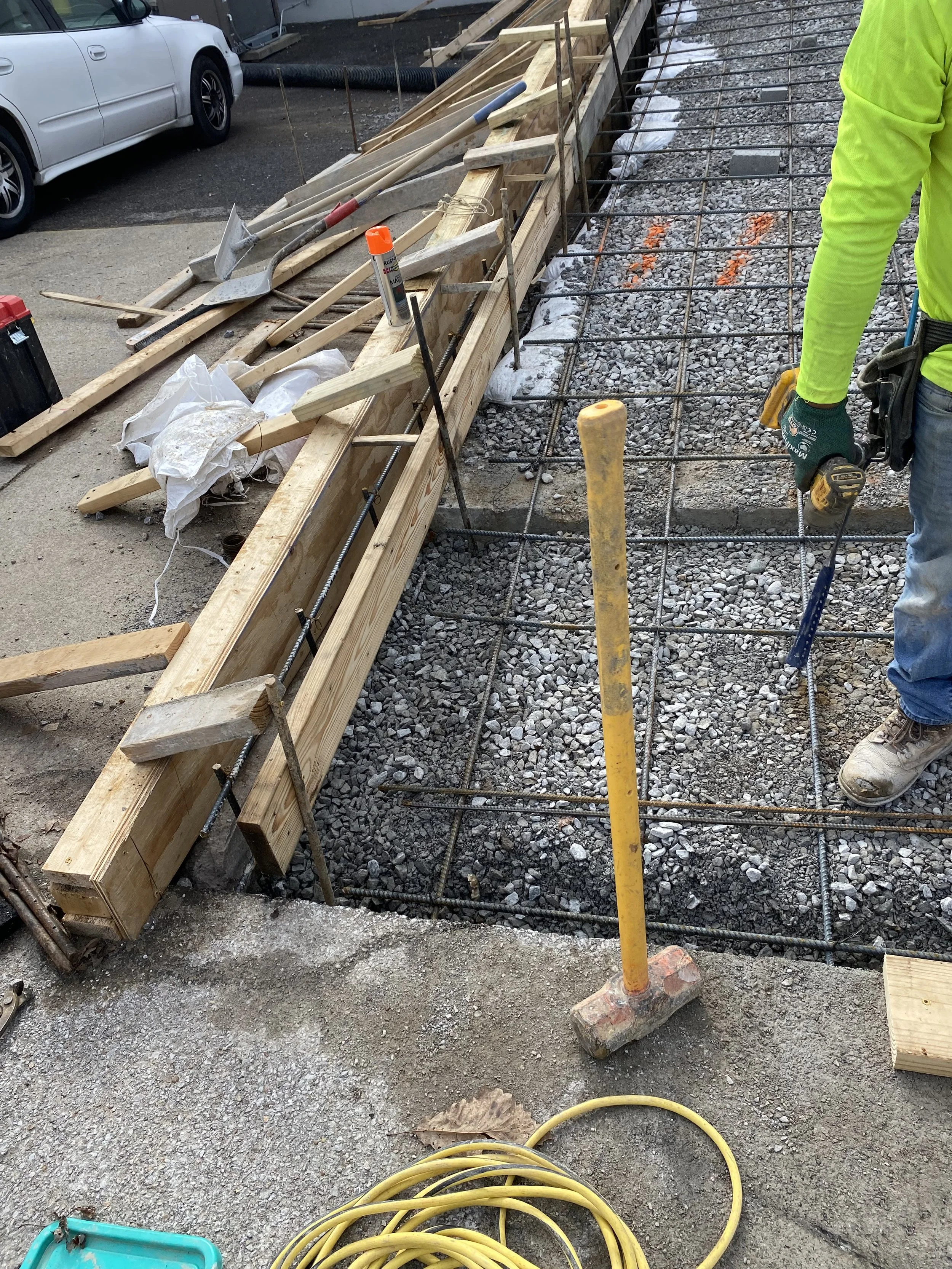 Construction site with rebar and wooden formwork for pouring foundation or sidewalk. Worker in high-visibility clothing with tools nearby.