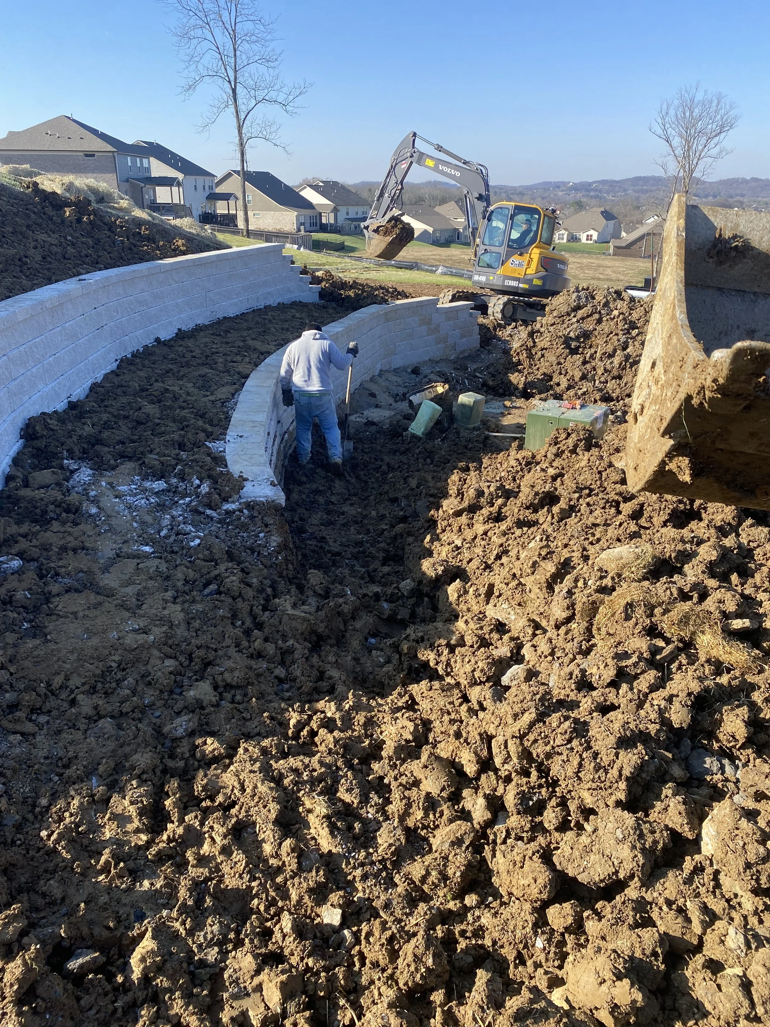 Construction site showing a worker installing a curved retaining wall made of concrete blocks, with an excavator nearby and several plastic containers on muddy ground.