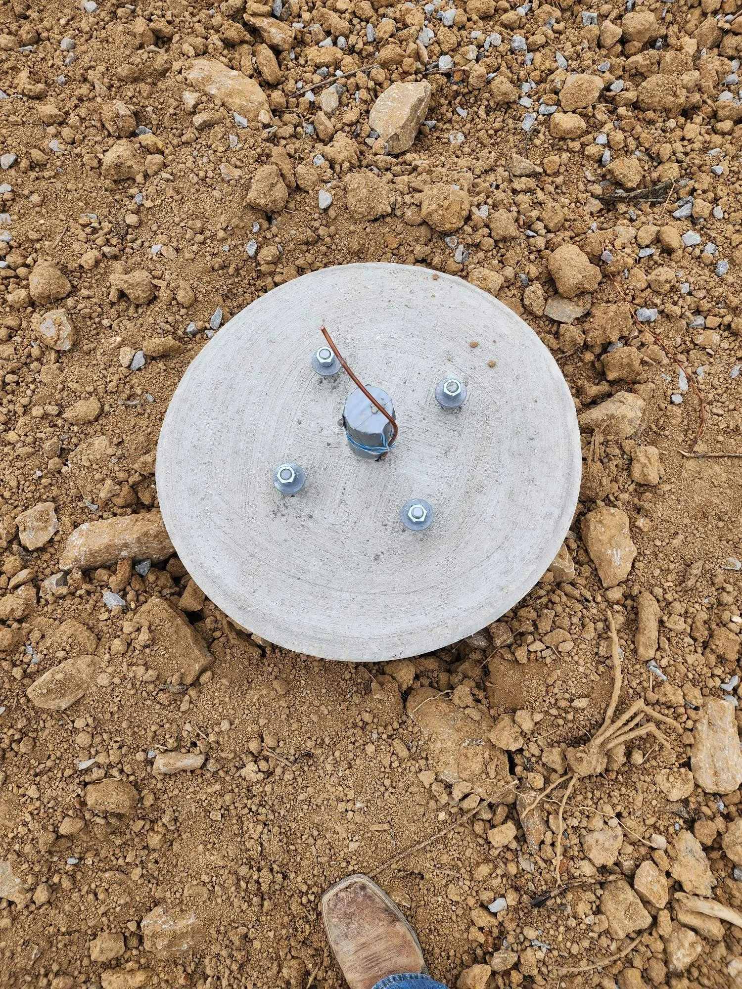 A concrete foundation with bolts and wires on dirt ground, with a human foot wearing a worn brown boot visible at the bottom of the image.