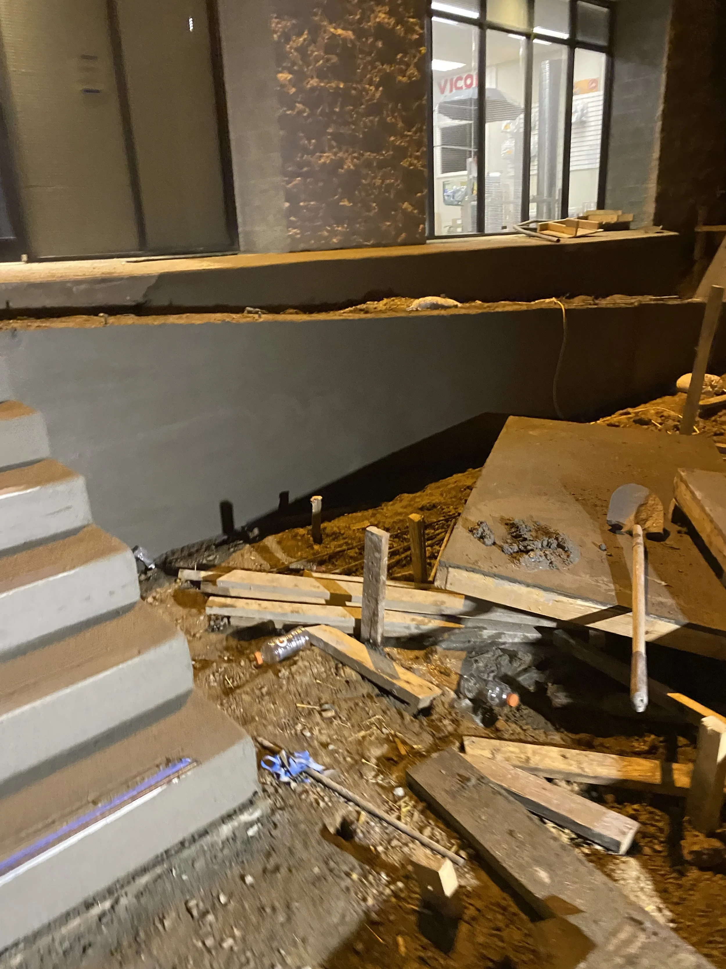 Construction site at night with exposed ground, scattered wooden planks, bottles, and construction tools in front of a building with large windows and a door.
