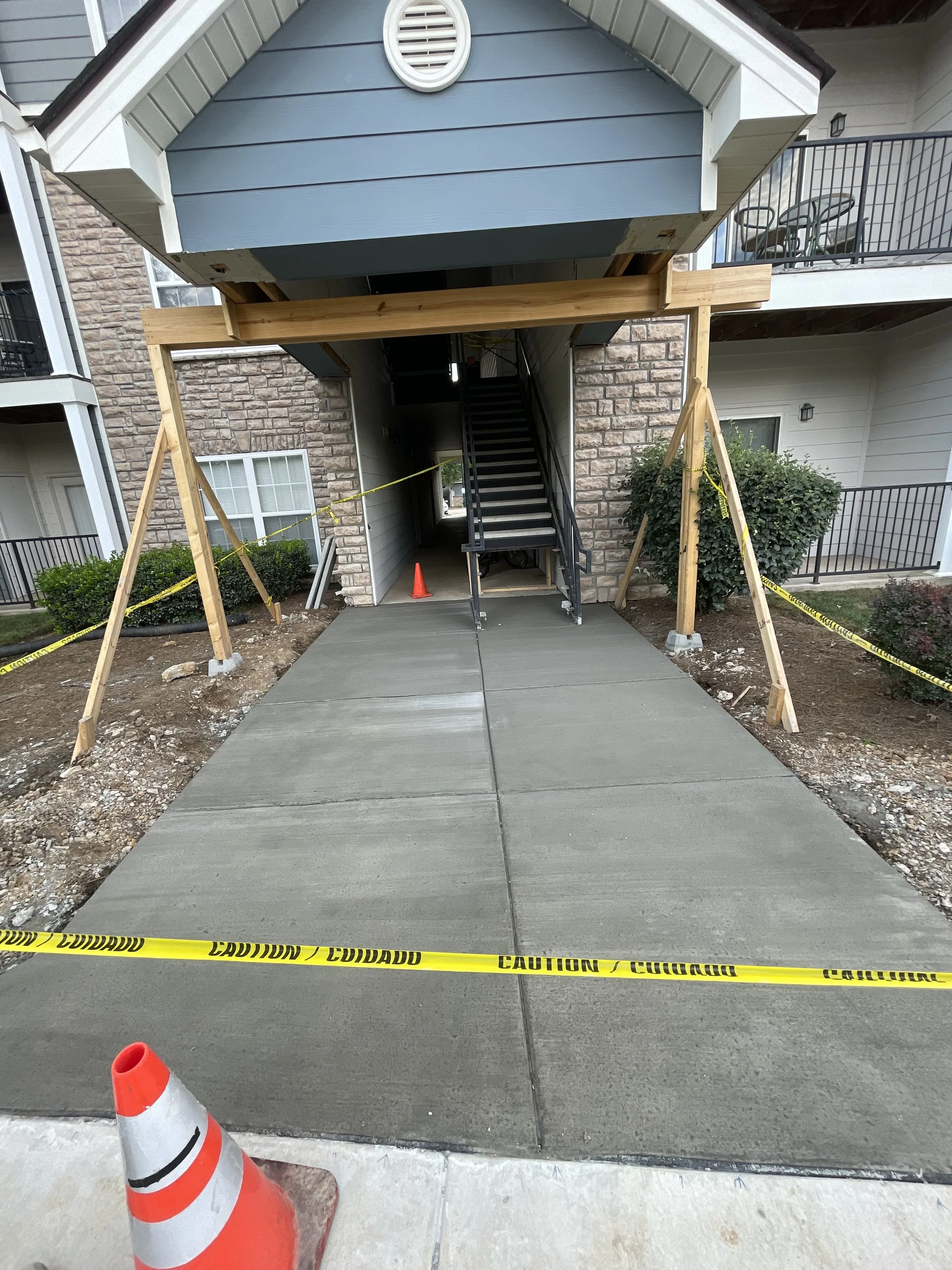 Construction site of a staircase and covered area at an apartment complex, with safety caution tape, orange traffic cone, and wooden supports.