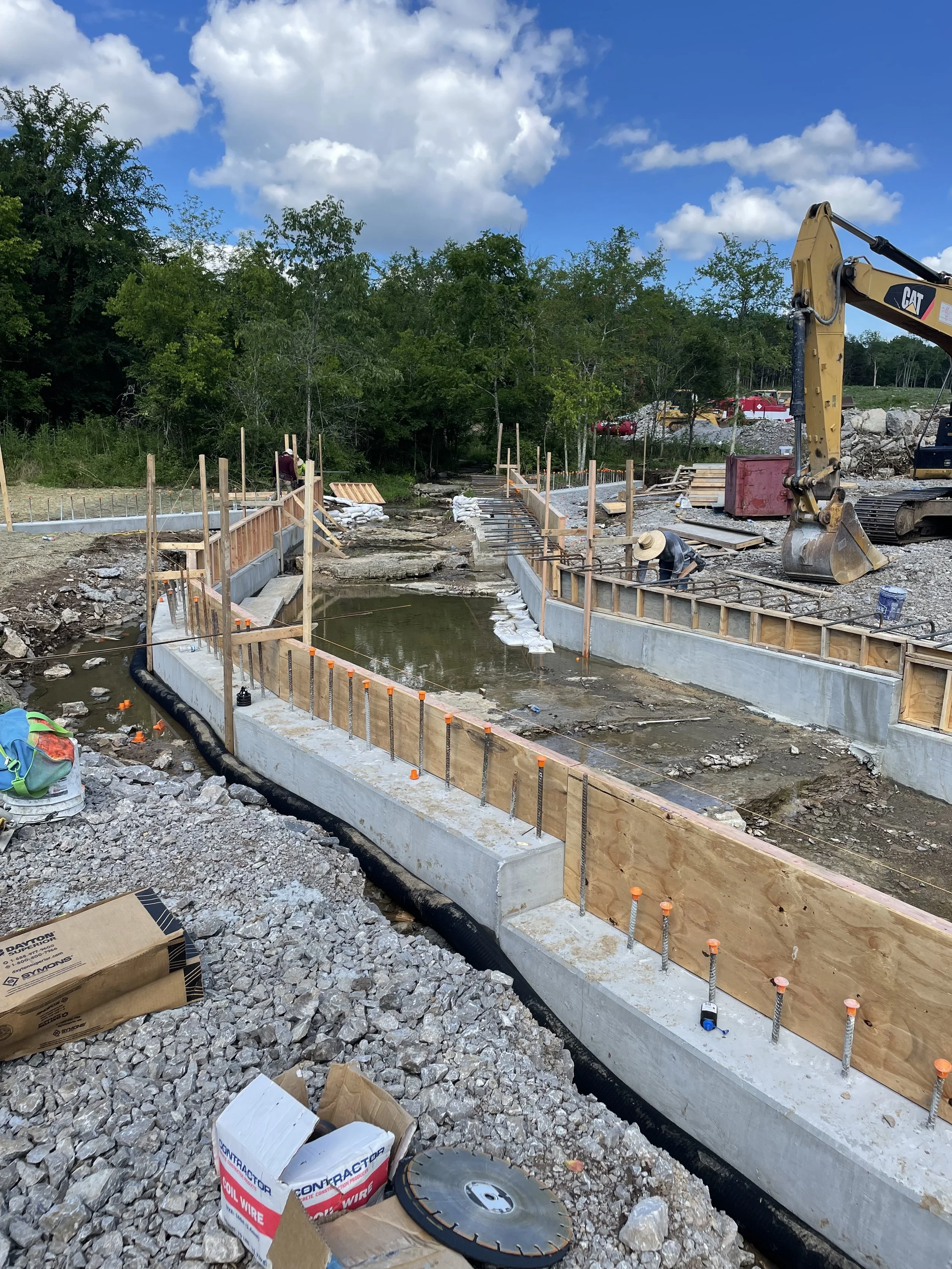 Construction site showing a curved concrete foundation and wooden formwork, with workers installing rebar and equipment like a small excavator, under a partly cloudy sky.