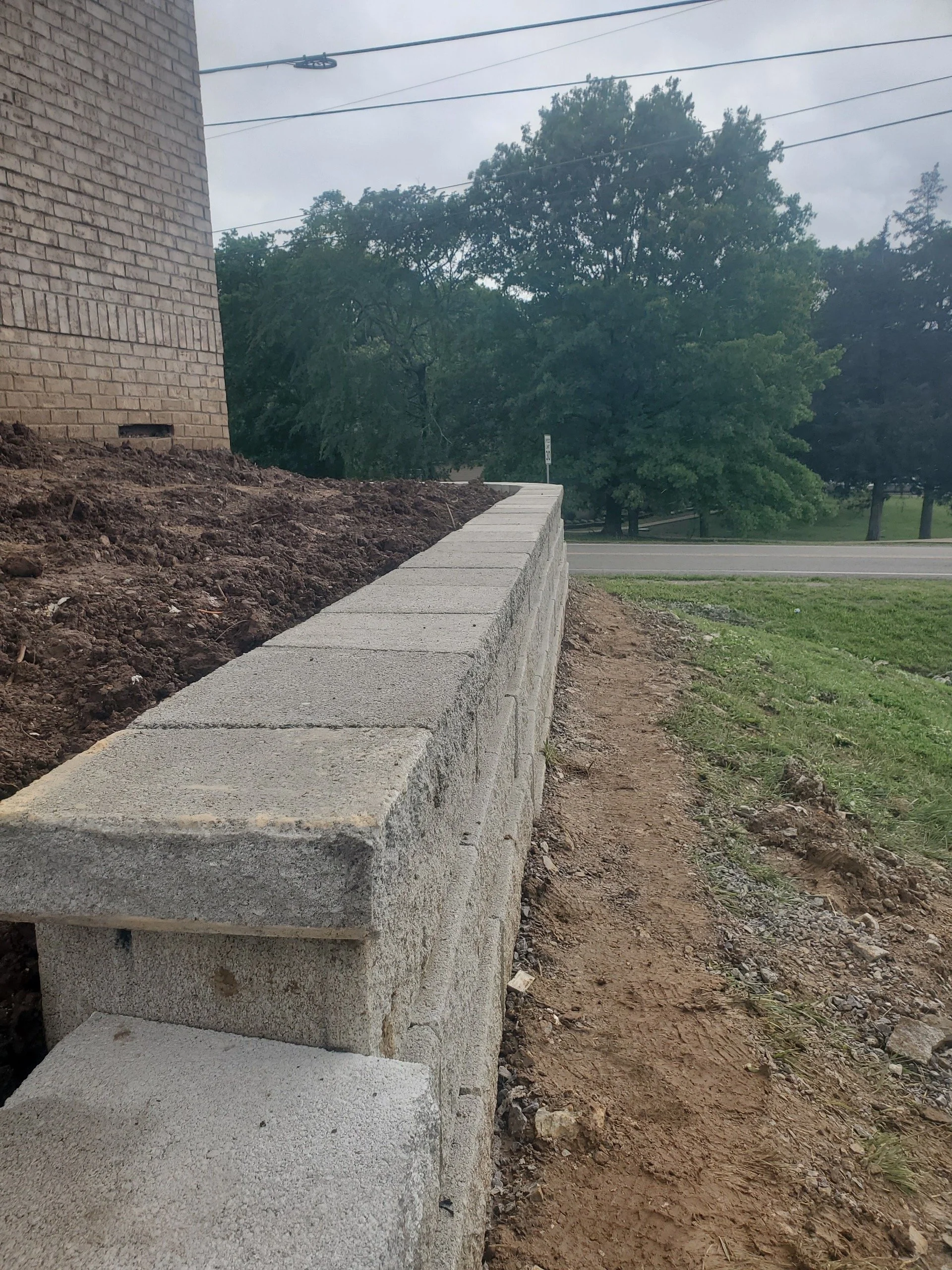 A new gray concrete block wall under construction next to a brick building, with a dirt pathway and green grass, and trees in the background under a cloudy sky.