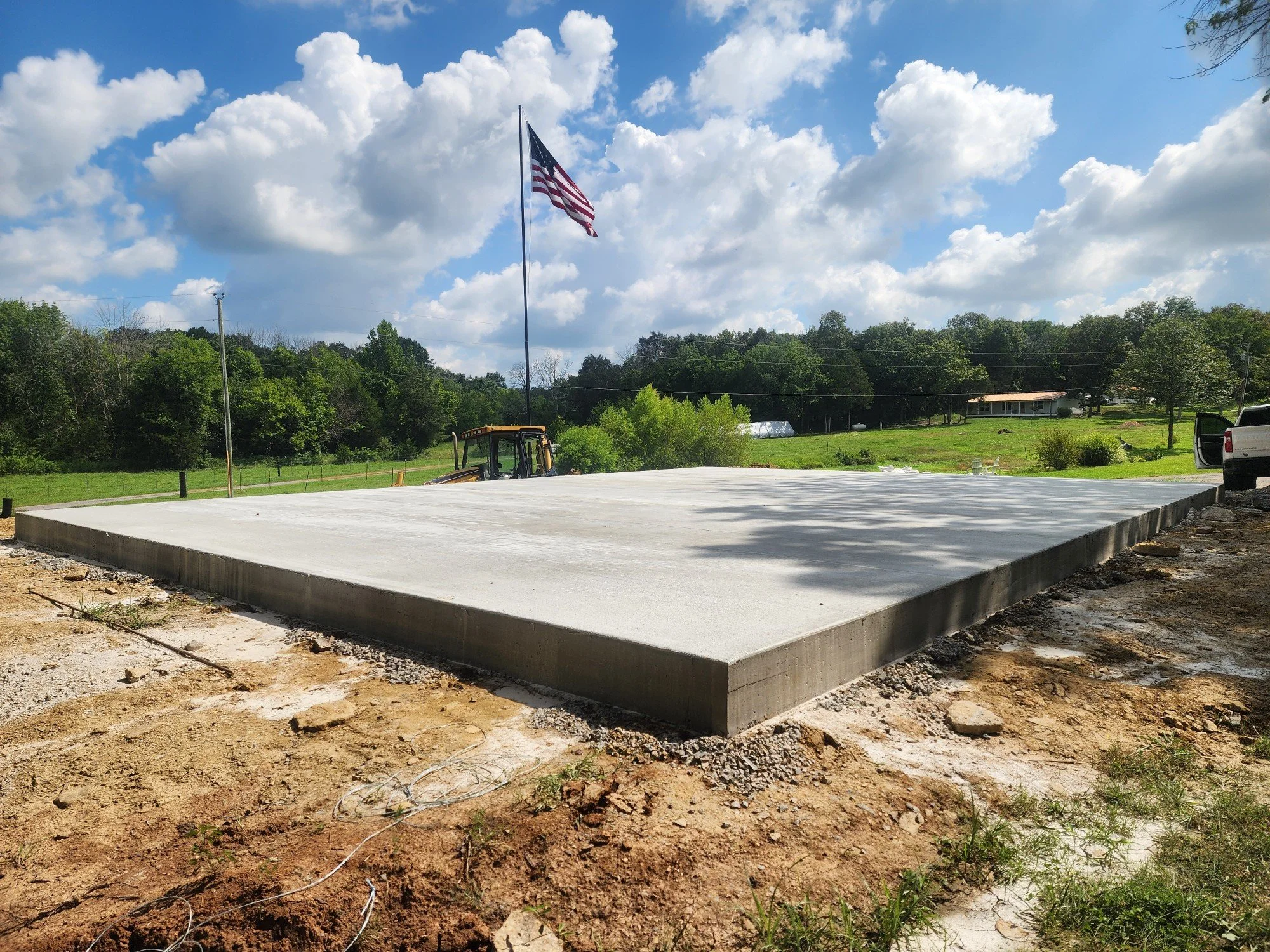 Freshly poured concrete slab foundation with an American flag flying on a pole, behind a construction vehicle, in outdoor rural area with green trees and houses in the background under a partly cloudy sky.