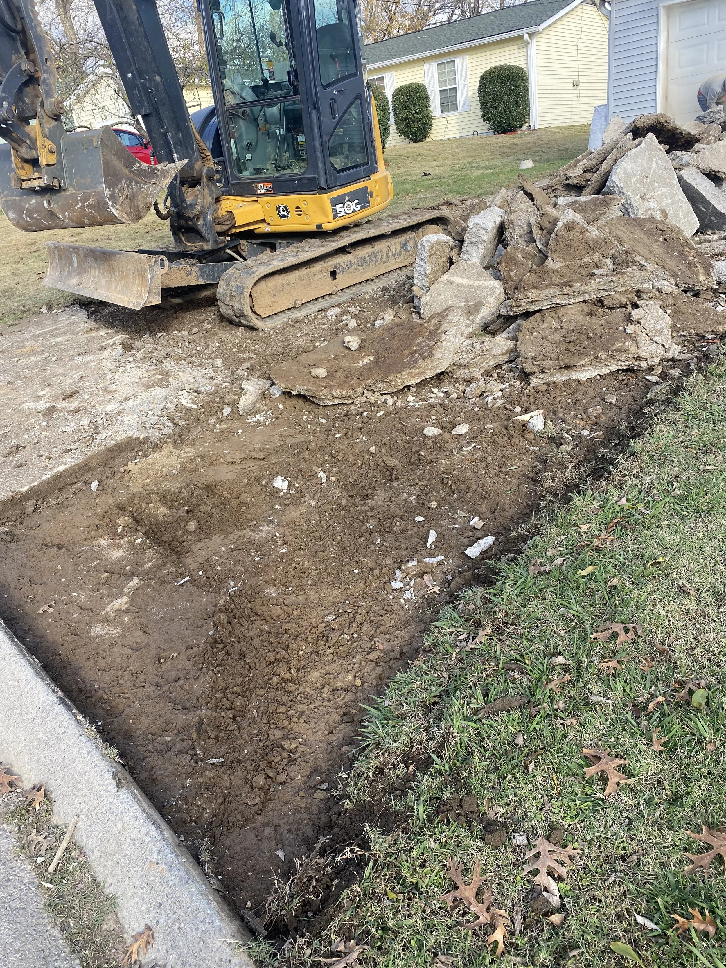 Excavation site next to a residential sidewalk with a yellow compact excavator and a pile of broken concrete and soil.