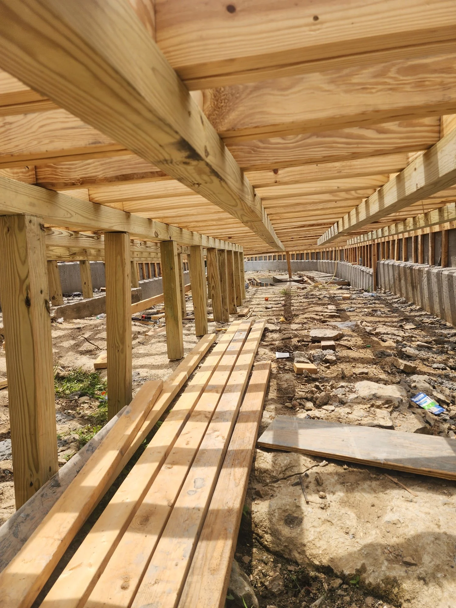 Underneath a wooden deck under construction, showing supporting wooden beams, planks, and a dirt and concrete foundation with scattered debris.