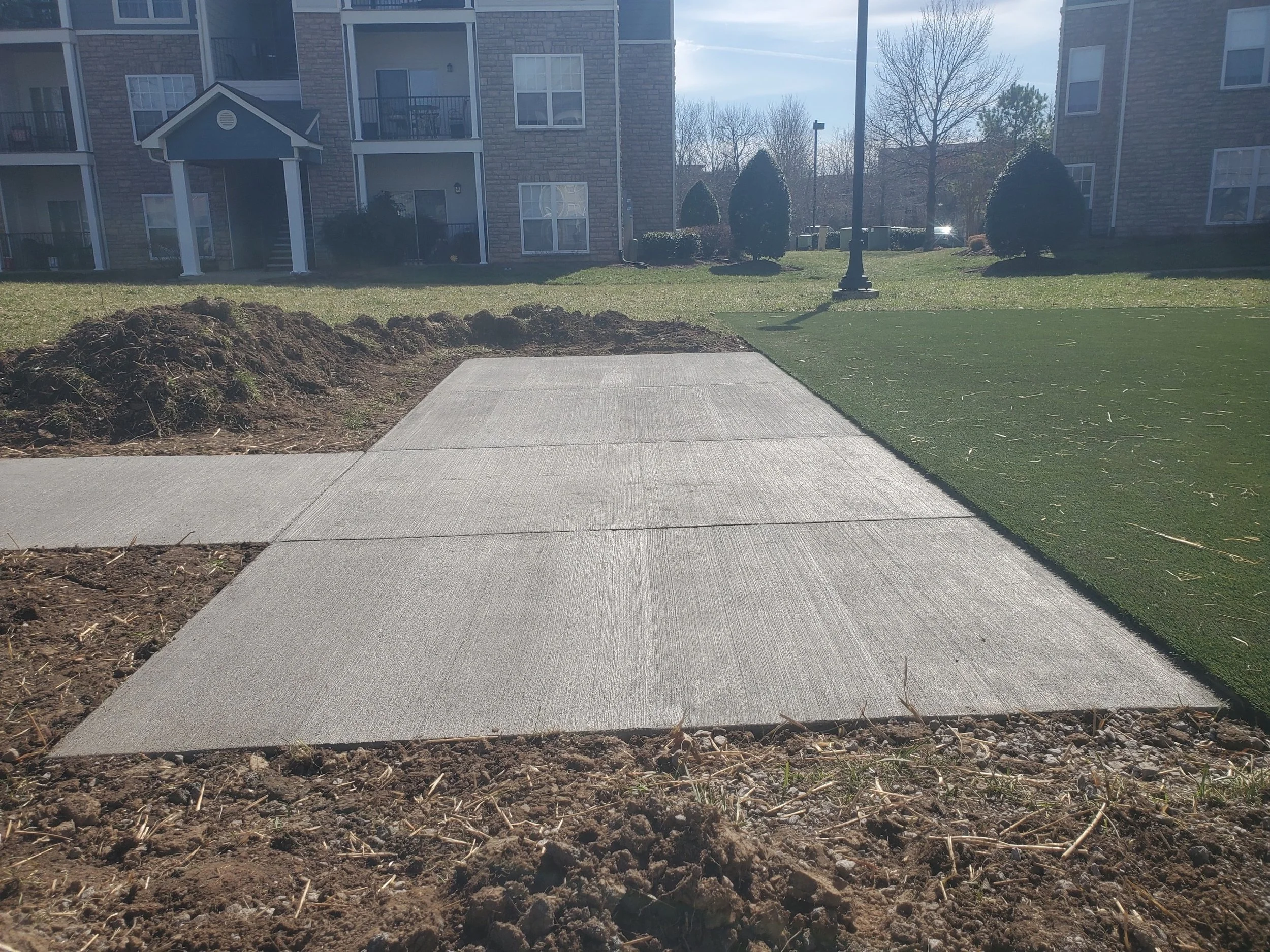 Freshly poured concrete sidewalk next to a patch of green artificial turf in an apartment complex courtyard.