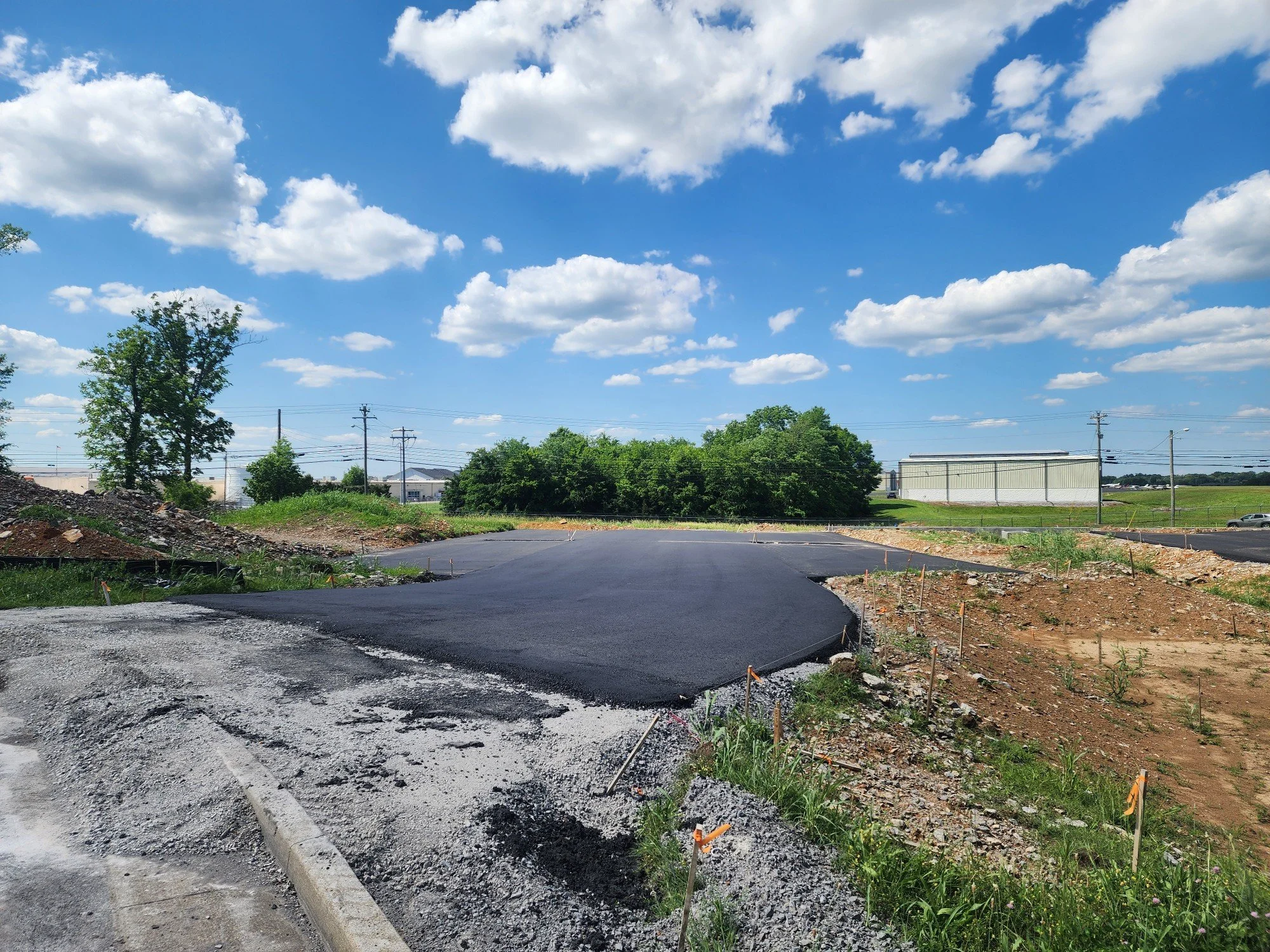 Newly paved and freshly asphalted road under a partly cloudy sky with green trees and construction markers on the side.