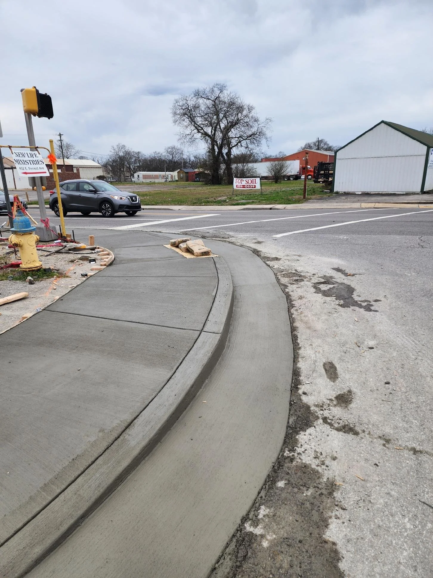 Construction site on a corner with fresh poured concrete sidewalk, construction materials, and a street with cars passing by. Signs advertising 'New Life Ministries' and 'Top Soil' are visible in the background.