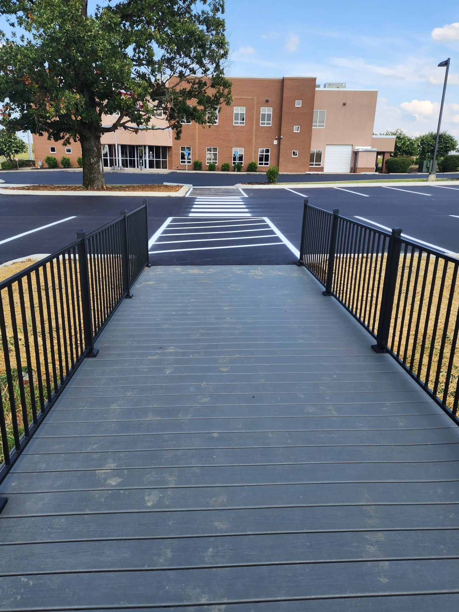 A small bridge with black metal railings leading over a parking lot to a sidewalk and a brick building with glass doors. There are trees and white parking line markings visible.