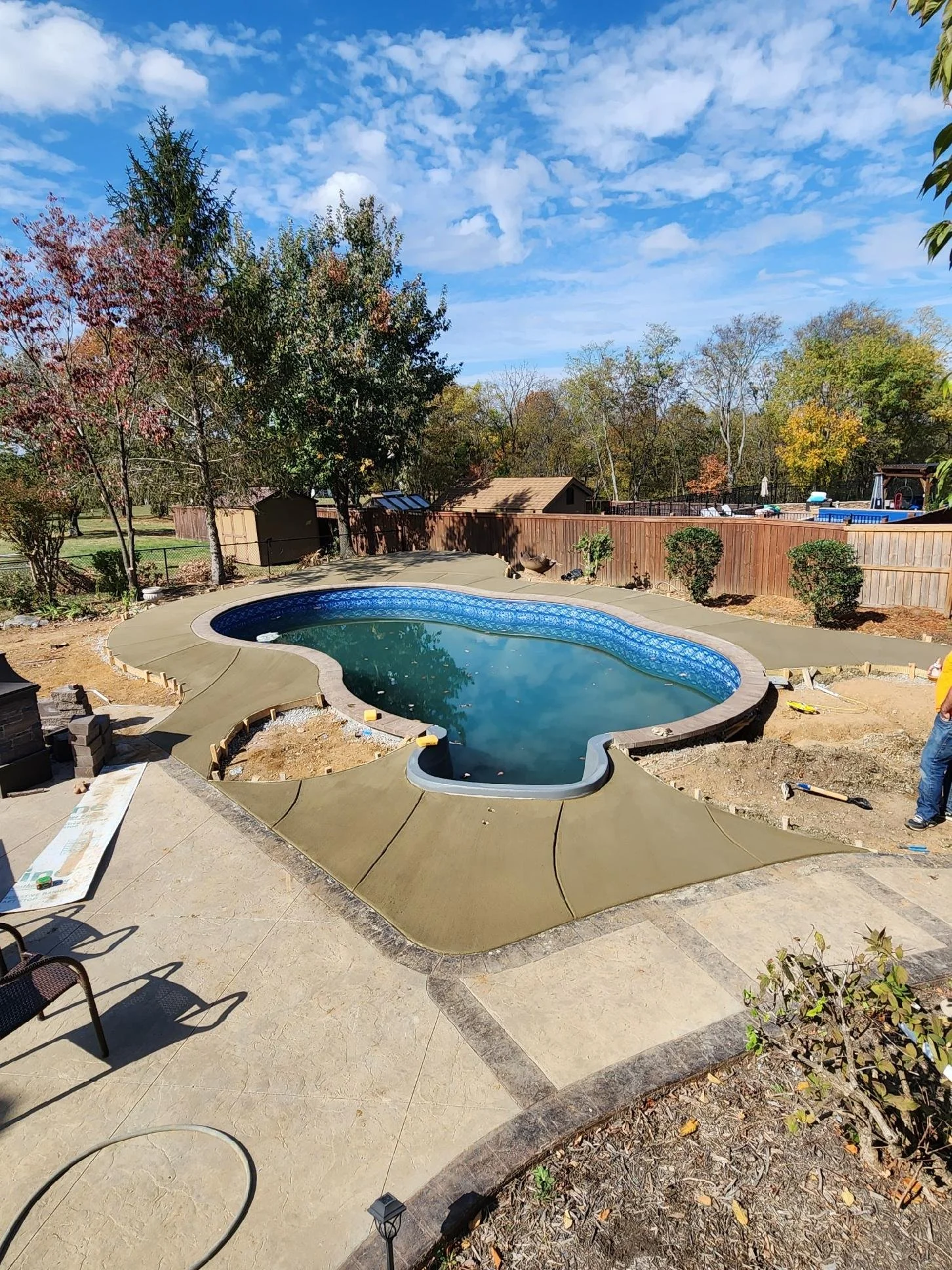 In-ground swimming pool under construction in backyard with surrounding concrete finish, trees, and a wooden fence, on a partly cloudy day.