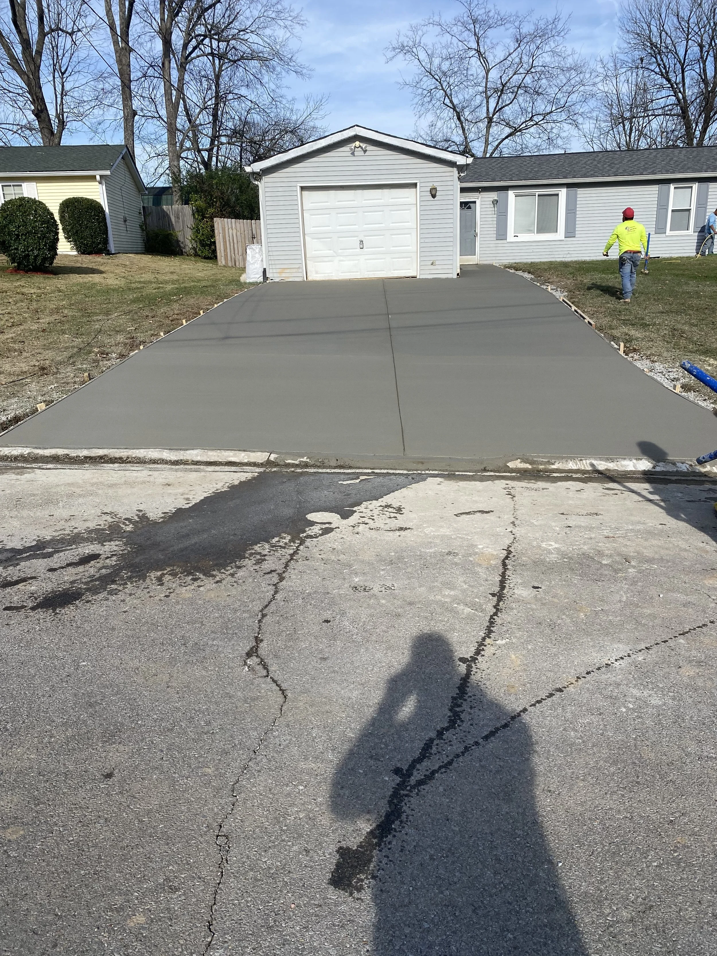 Freshly poured concrete driveway leading to a white garage in a residential yard with two workers nearby and trees in the background.