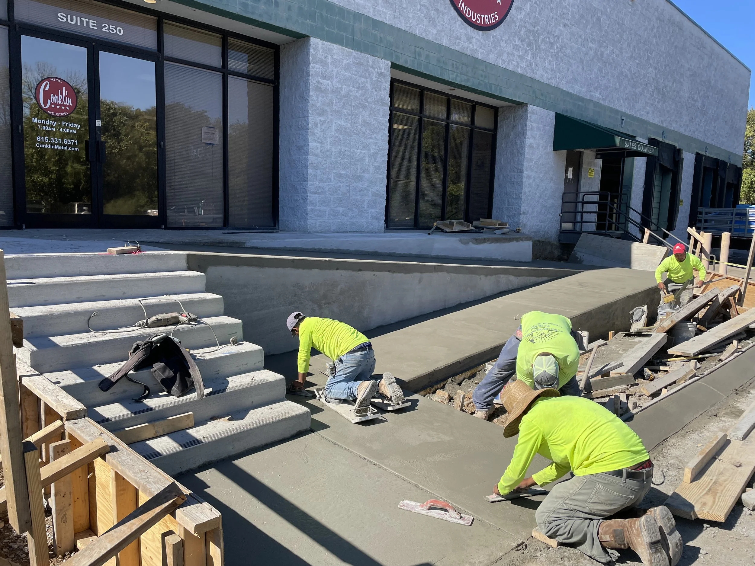 Construction workers in bright yellow shirts and hats pouring and smoothing concrete for an outdoor sidewalk or ramp in front of commercial buildings, with stairs and a ramp visible.