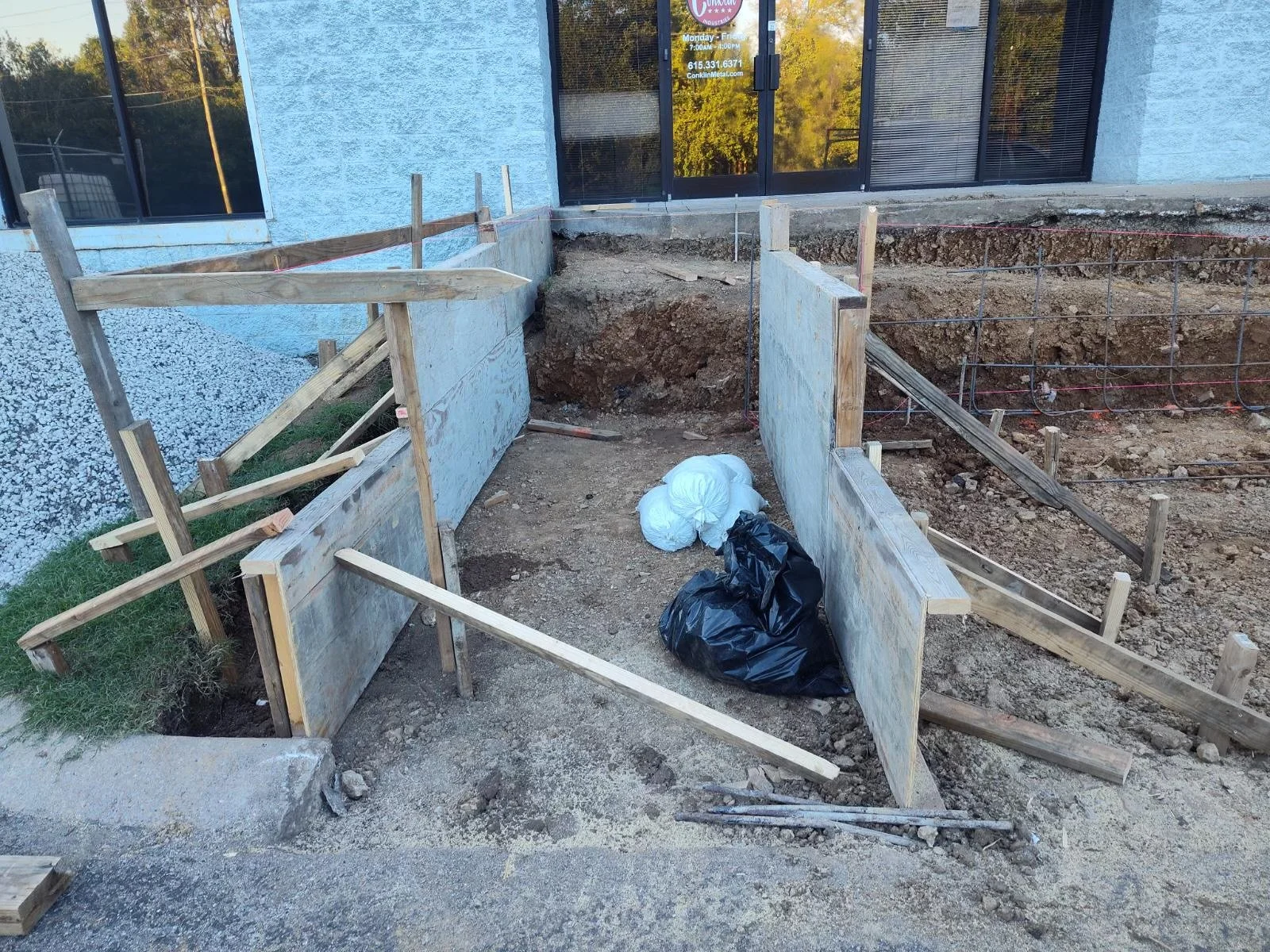 Construction site with dirt walls and wooden support panels, bags of trash, and some scattered wood pieces, next to a building with a blue exterior and large windows.