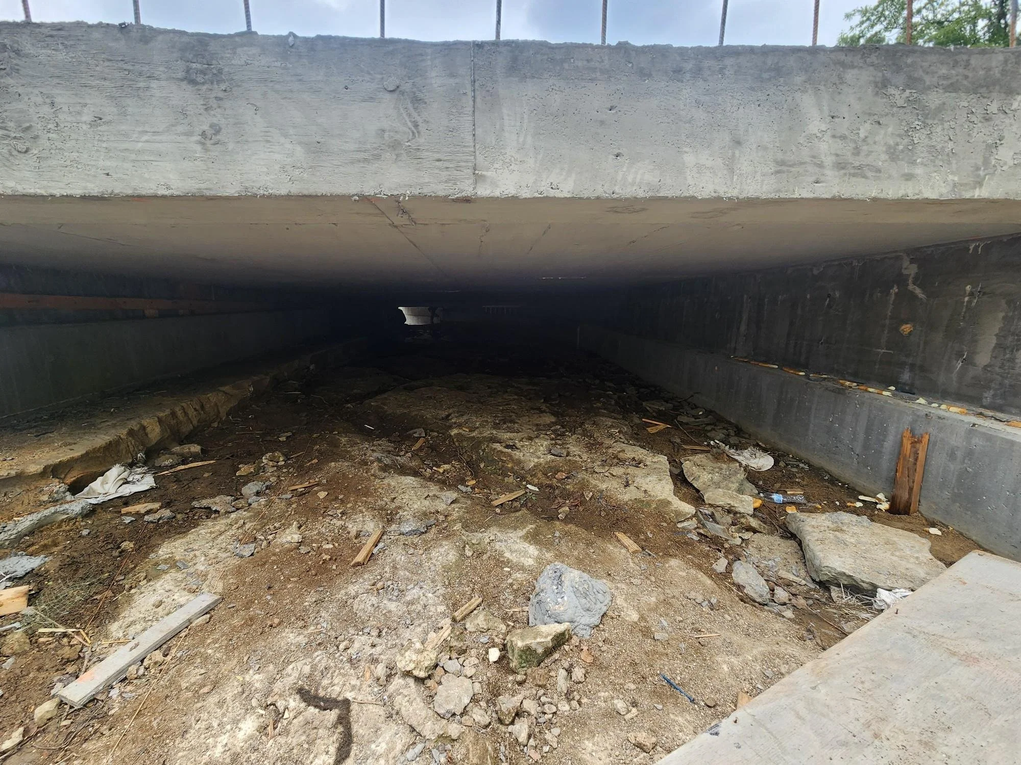 Underneath a concrete bridge or overpass with dirt, rocks, and debris on the ground.