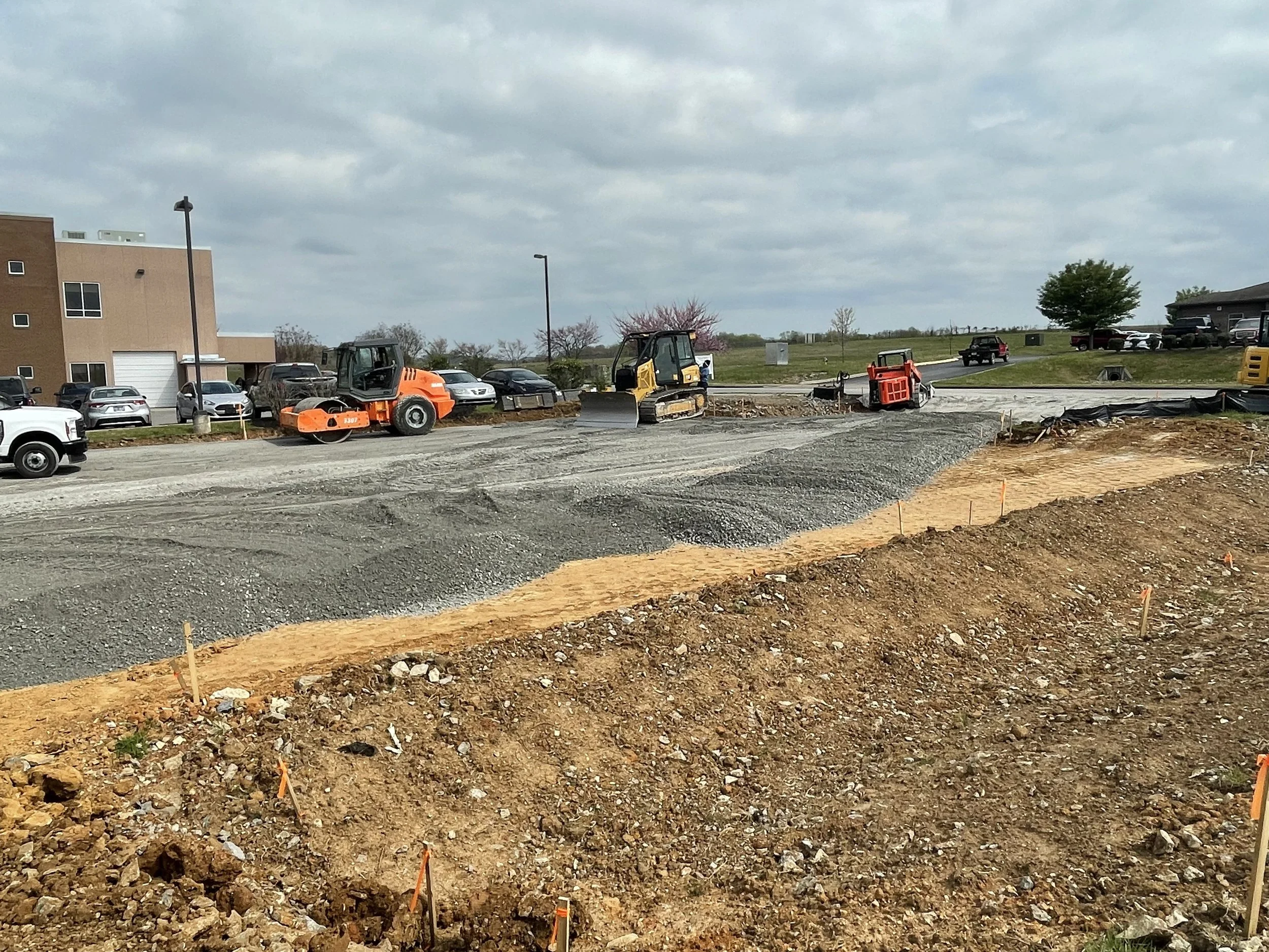 Construction site with dirt and gravel being prepared for paving, construction equipment including a roller and compactors, and parked cars in the background.