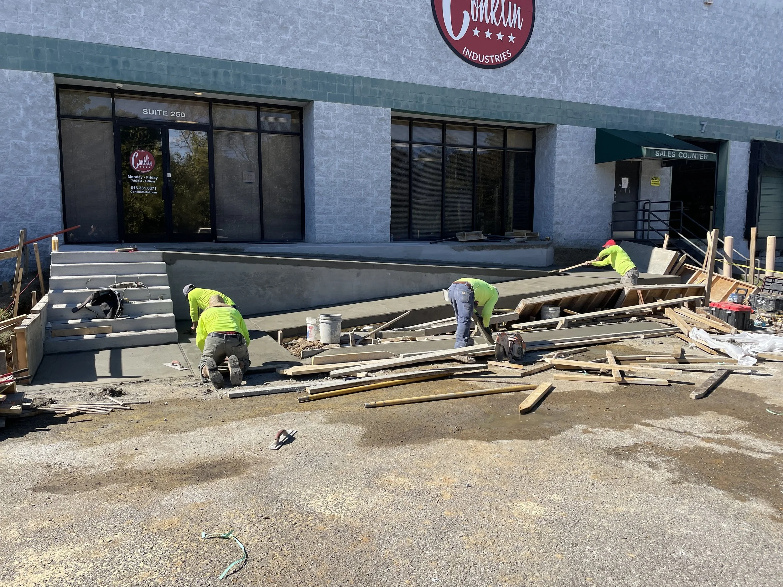 Construction workers pouring concrete on a sidewalk outside a building with a sign reading 'Cream Industries' and a glass door labeled 'Suite 250'.