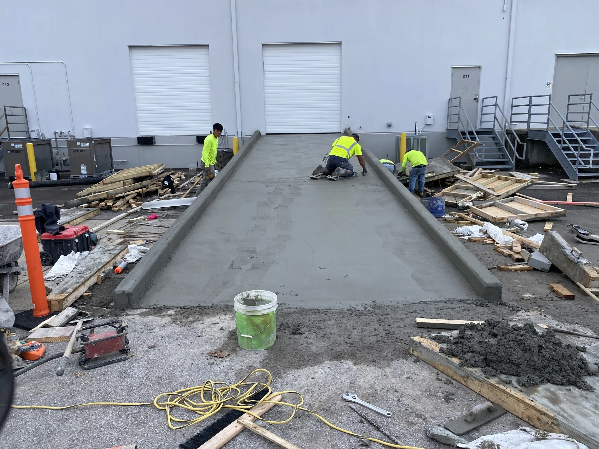 Construction workers pouring and smoothing concrete on a new sidewalk or ramp outside a warehouse, with construction tools and materials scattered around.