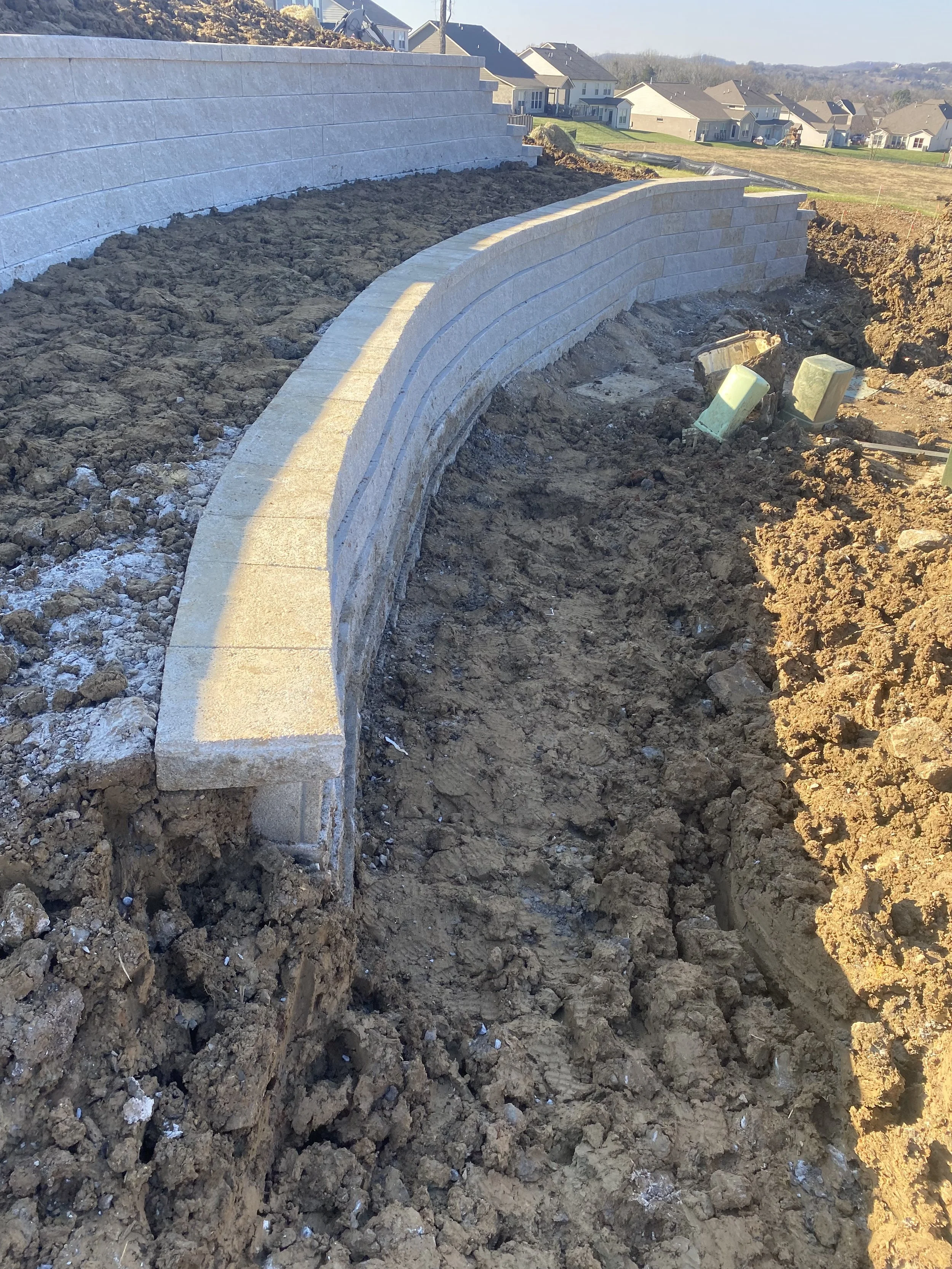 Construction site with a curved stone retaining wall being built on a dirt lot, with residential houses in the background.
