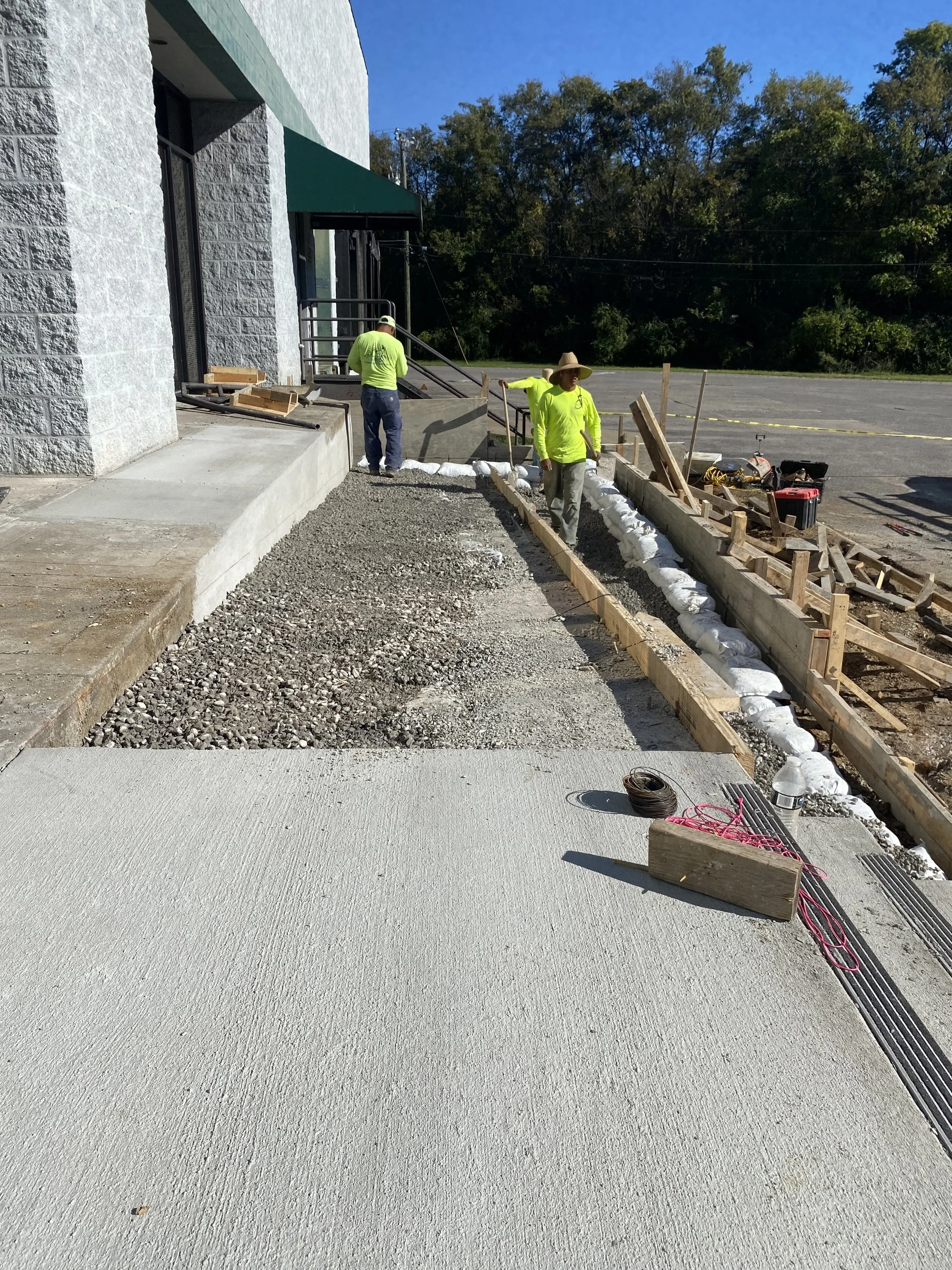 Construction workers laying concrete sidewalk outside a building with a blue sky and trees in the background.