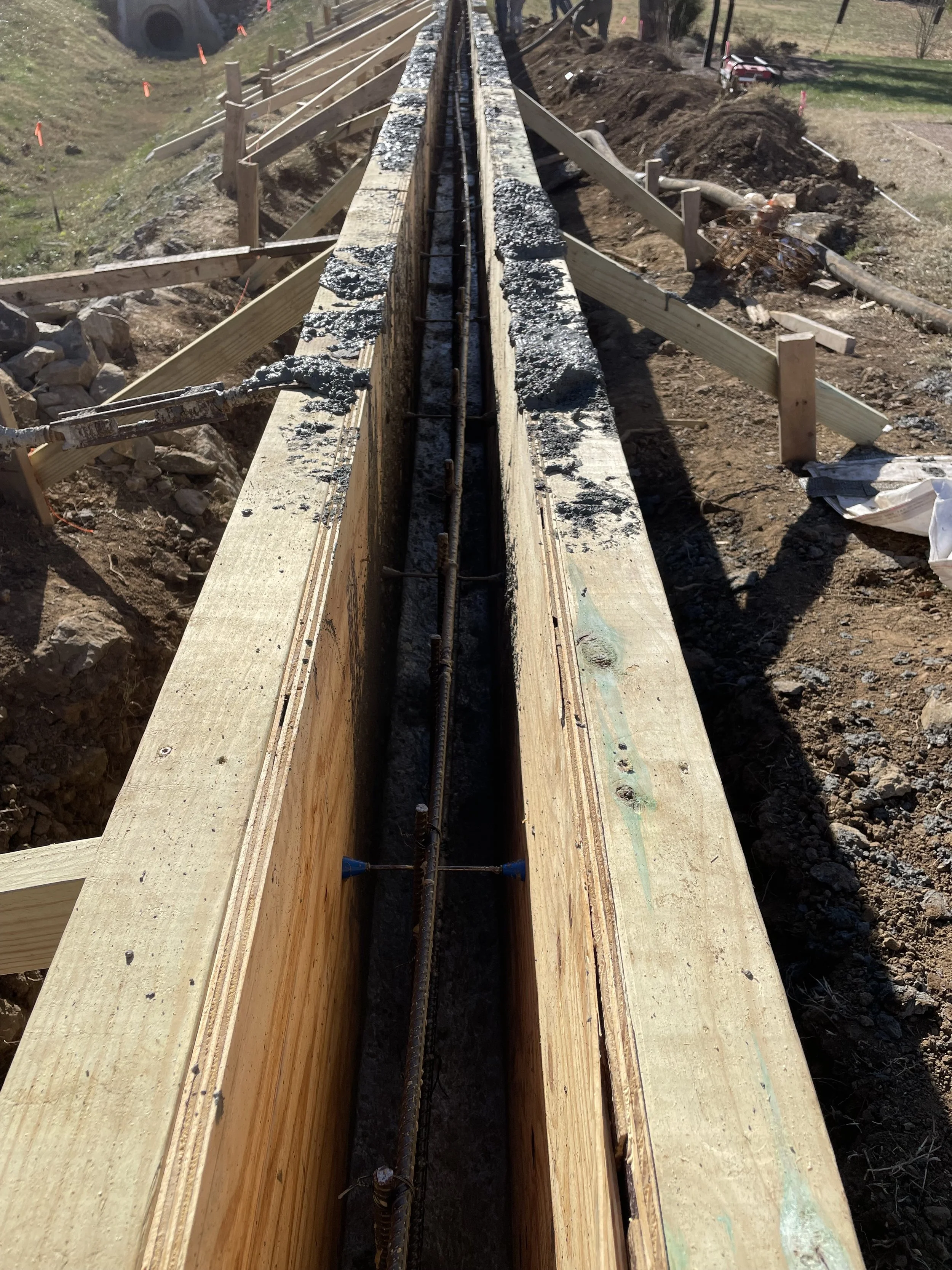 Construction site showing a wooden form for a concrete foundation with rebar inside, surrounded by dirt and construction tools.