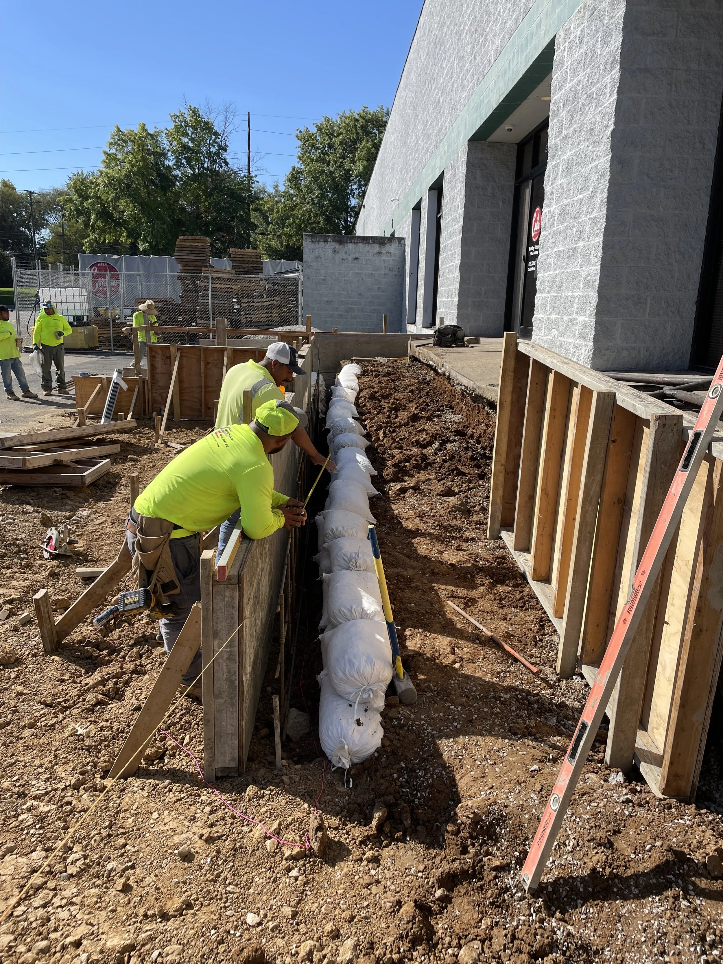 Construction workers working on a foundation or trench outside a building, with some workers measuring.