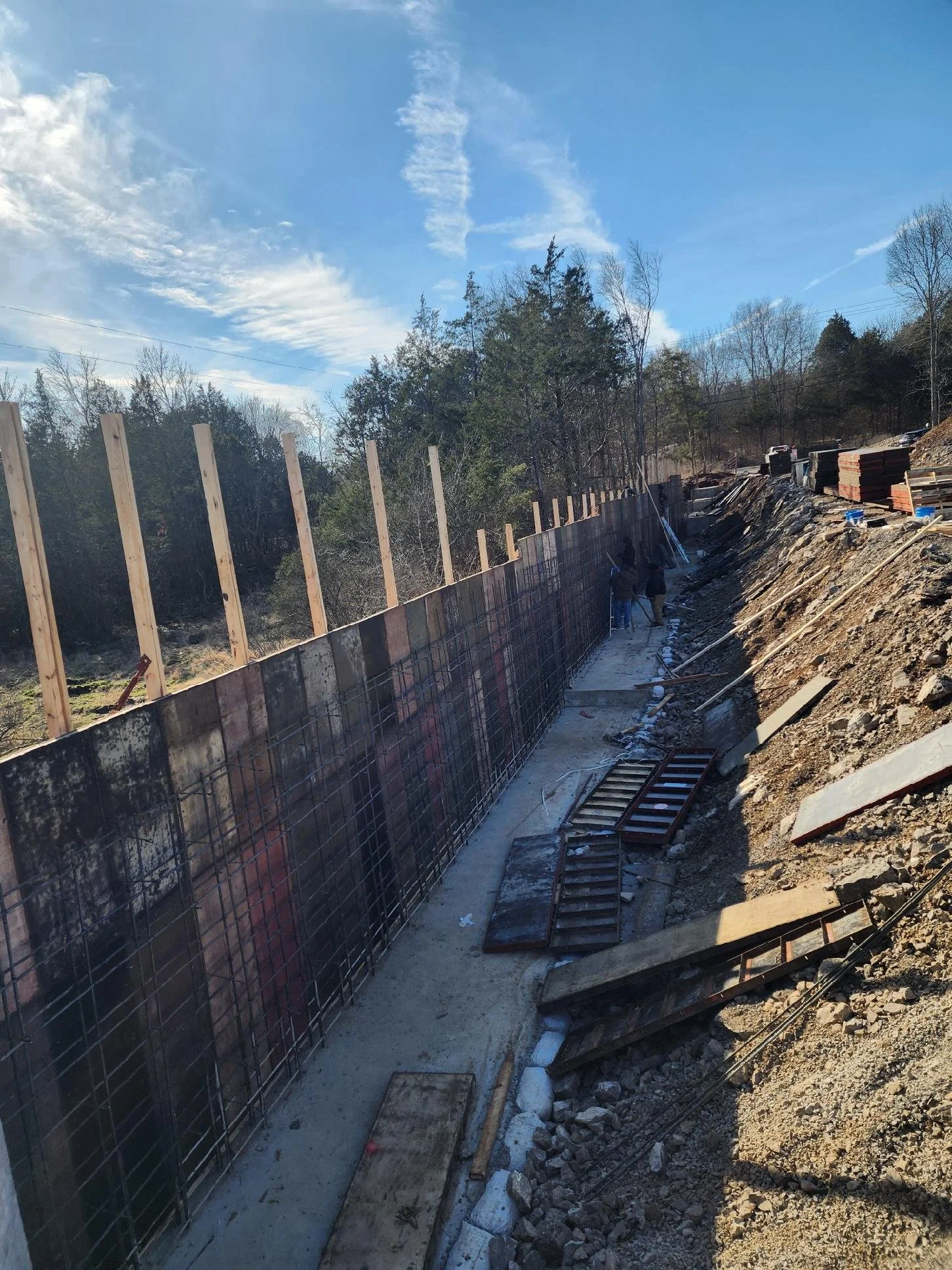 Construction site with partially built retaining wall, wooden support beams, and workers installing reinforcement steel within a trench.