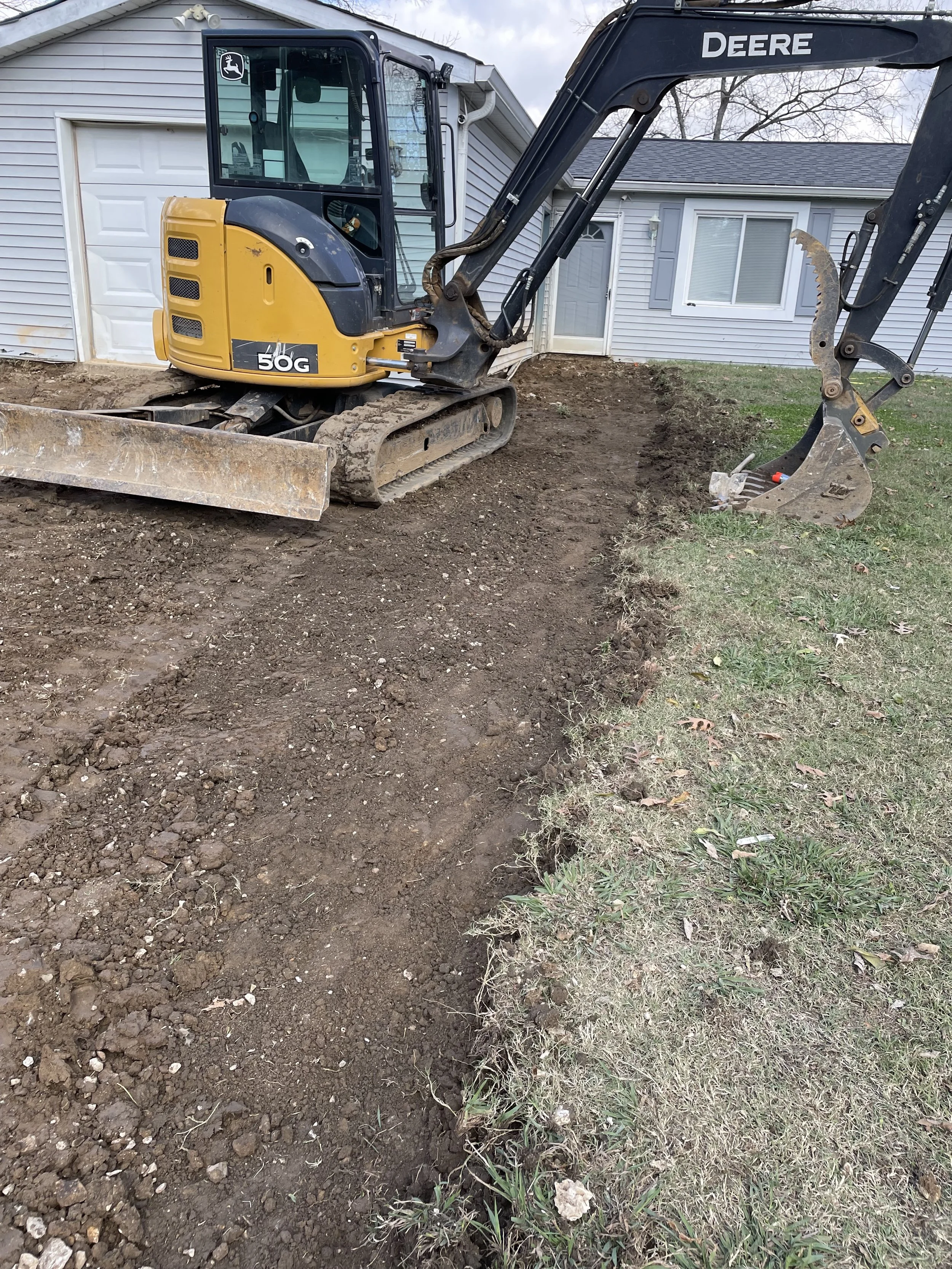 A small excavator working on digging a trench in a grassy yard near a house with siding and a garage door.