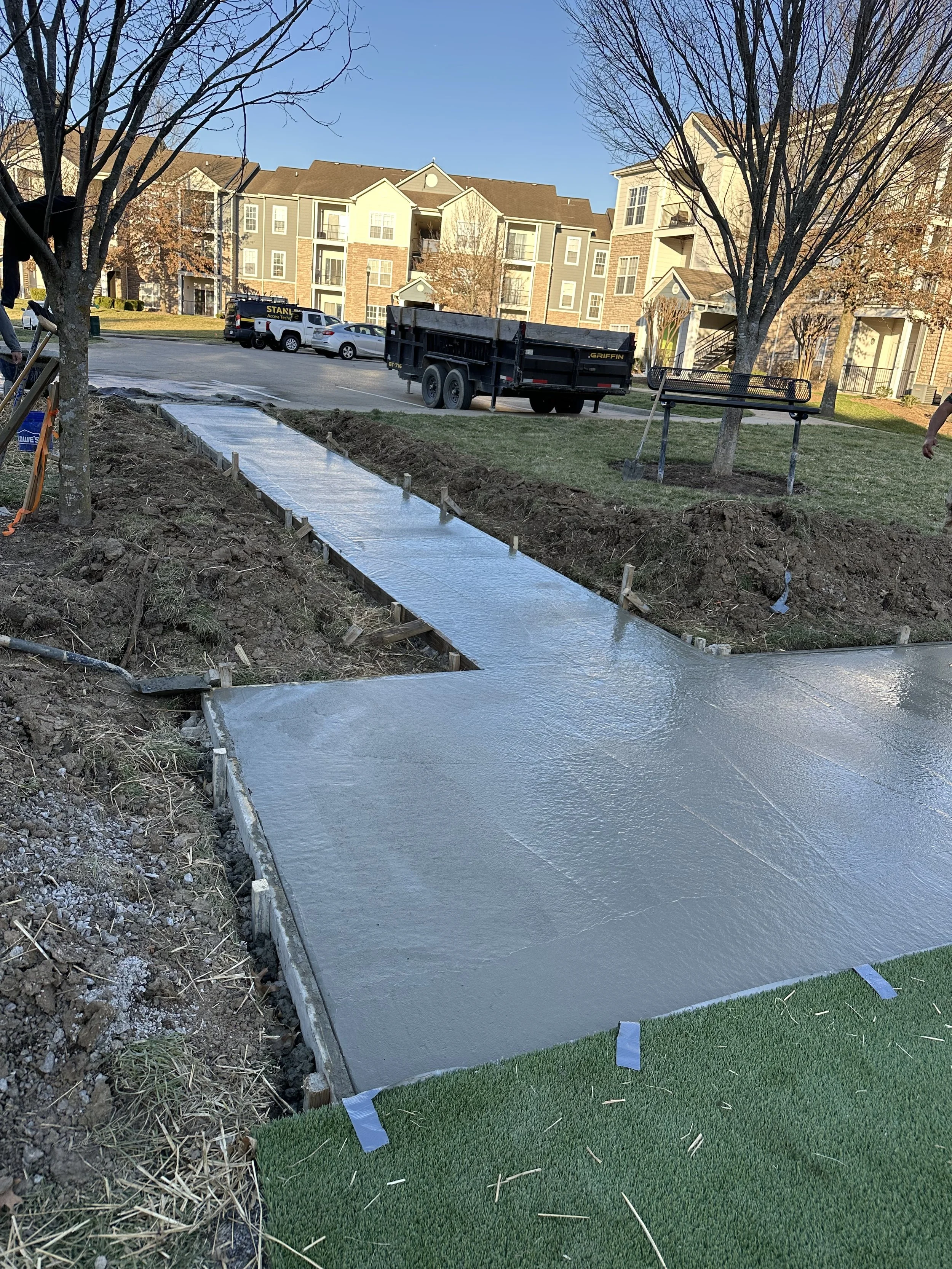 Concrete sidewalk under construction in a residential neighborhood with multi-story apartment buildings, leafless trees, parked cars, and a blue sky.