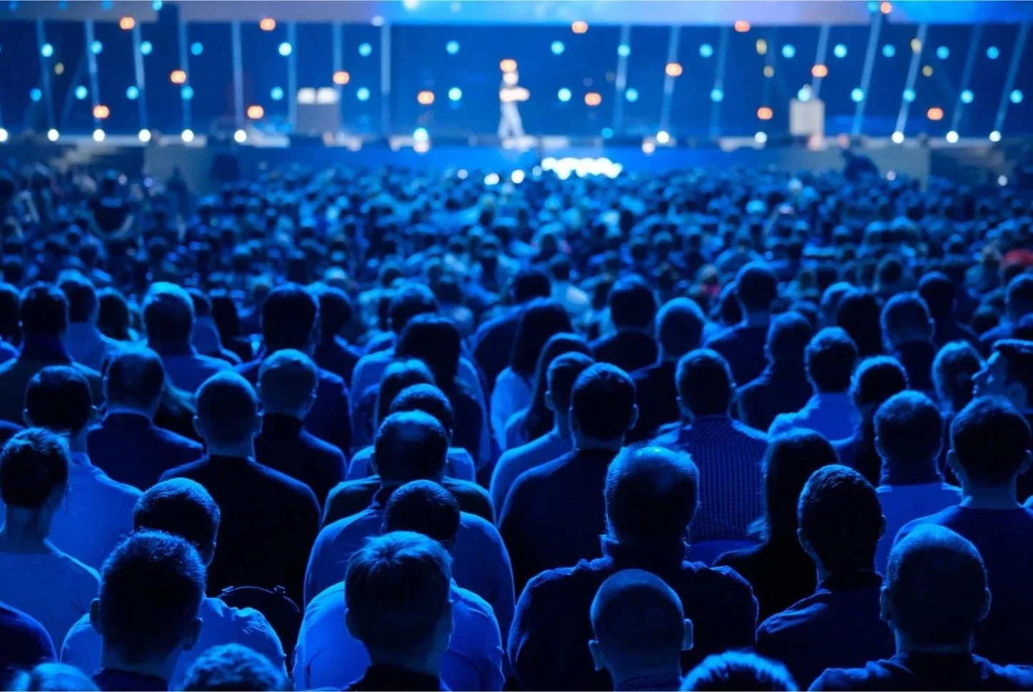 A large audience seated facing a stage with two people speaking, illuminated with blue stage lights.