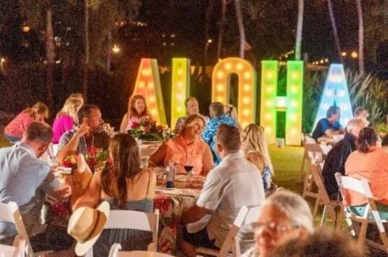 Outdoor gathering at night with people sitting at tables, in front of illuminated large letters spelling 'ALOHA' with a tropical background.