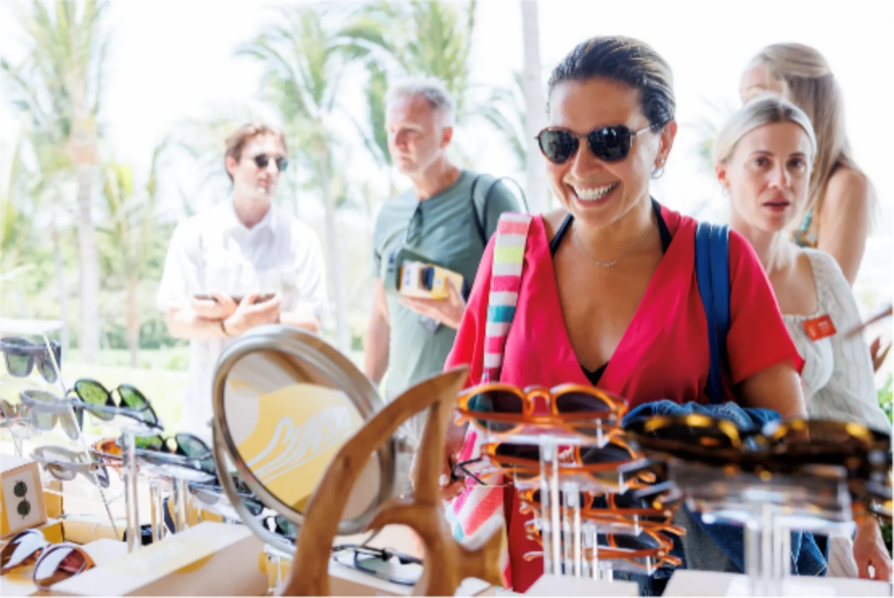 Smiling woman in sunglasses shopping for sunglasses at an outdoor market with other customers in background.