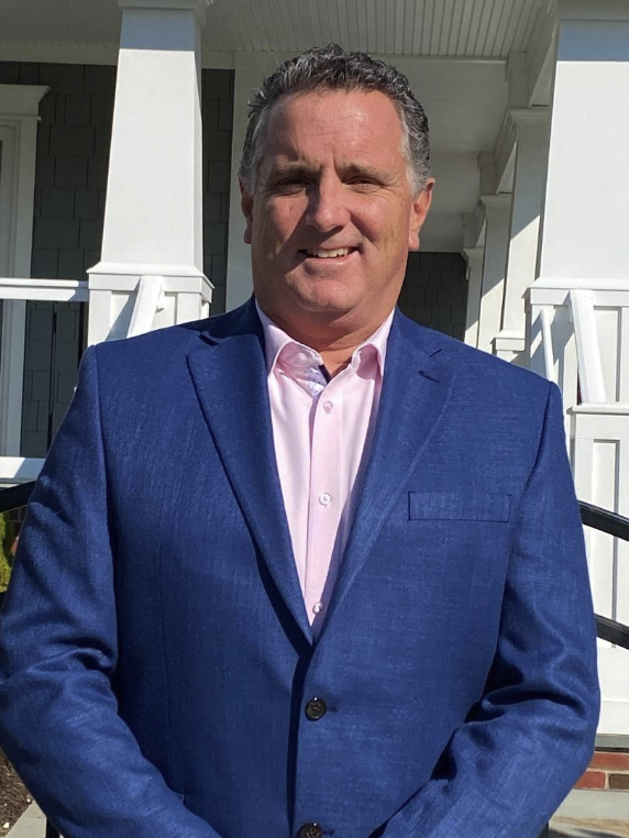 Man in blue suit and pink shirt standing outside in front of a house with white porch railings.