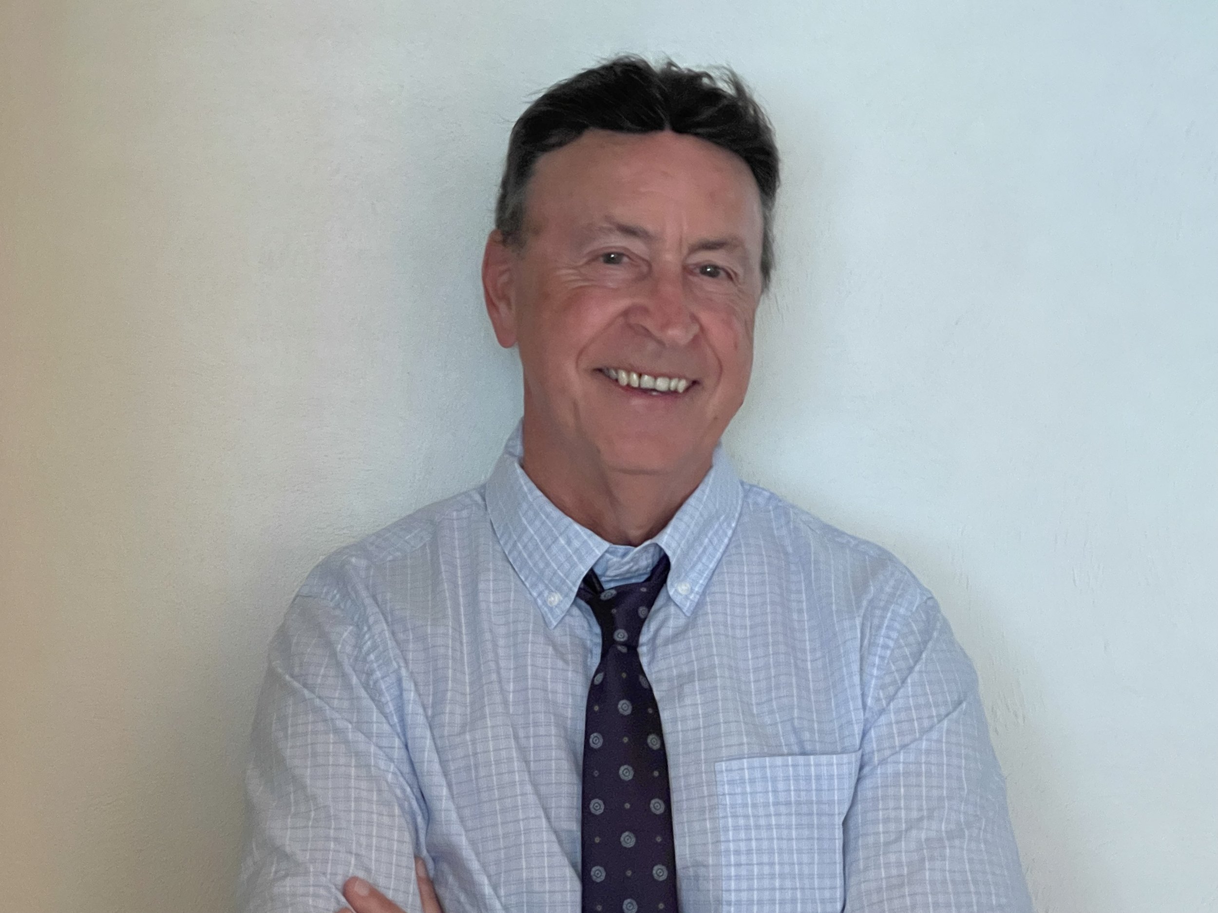A middle-aged man with dark hair smiling, wearing a light blue striped shirt and a dark tie, standing against a light-colored wall.