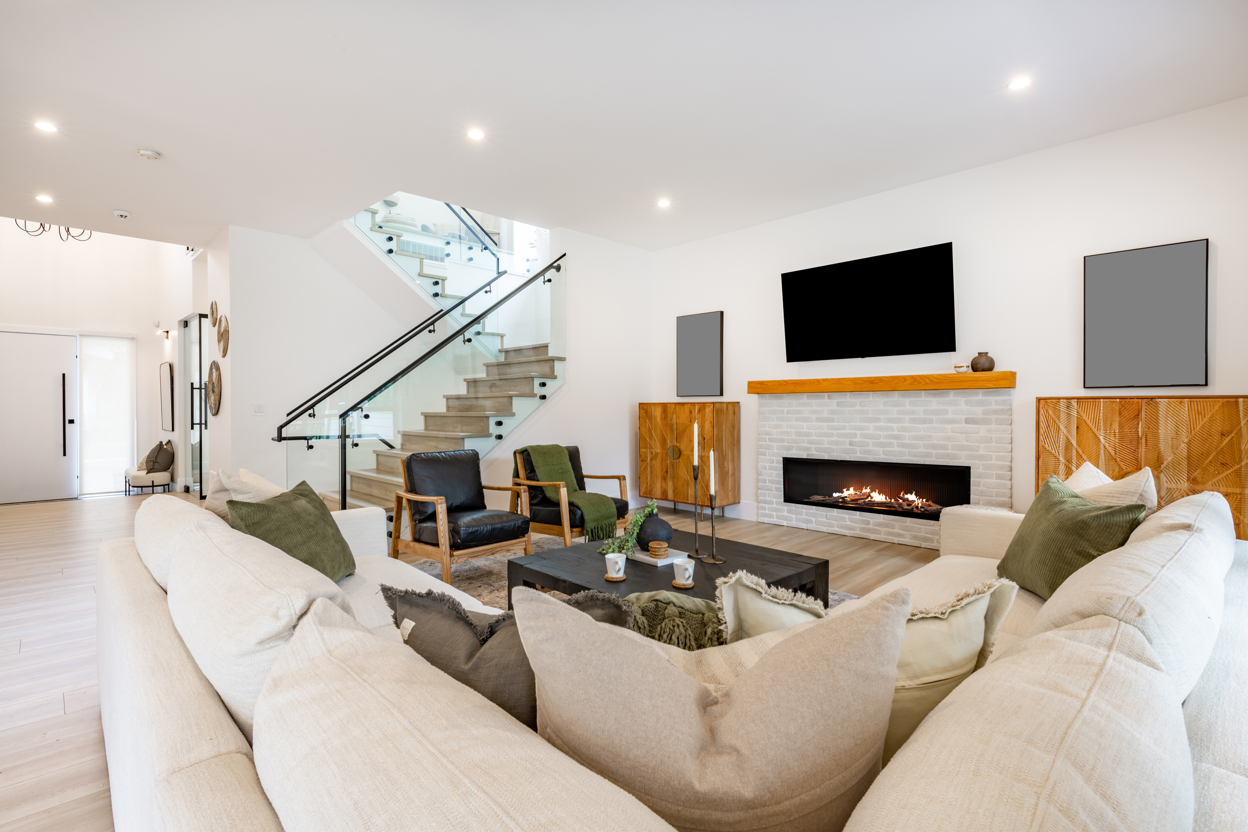 Modern living room featuring a white sectional sofa with green and beige pillows, a black coffee table with cups and a plant, a fireplace, and wall-mounted TV, with a staircase in the background.