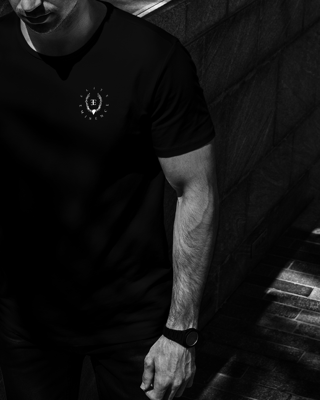 Close-up of a man wearing a black T-shirt with a small circular embroidered logo on the chest, standing against a textured wall with stairs nearby.