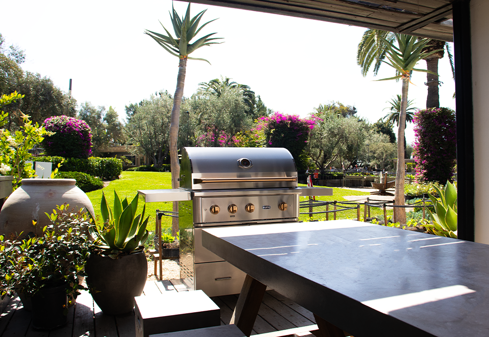 Outdoor patio with a stainless steel grill, potted plants, and a view of a lush garden with trees and flowering bushes.