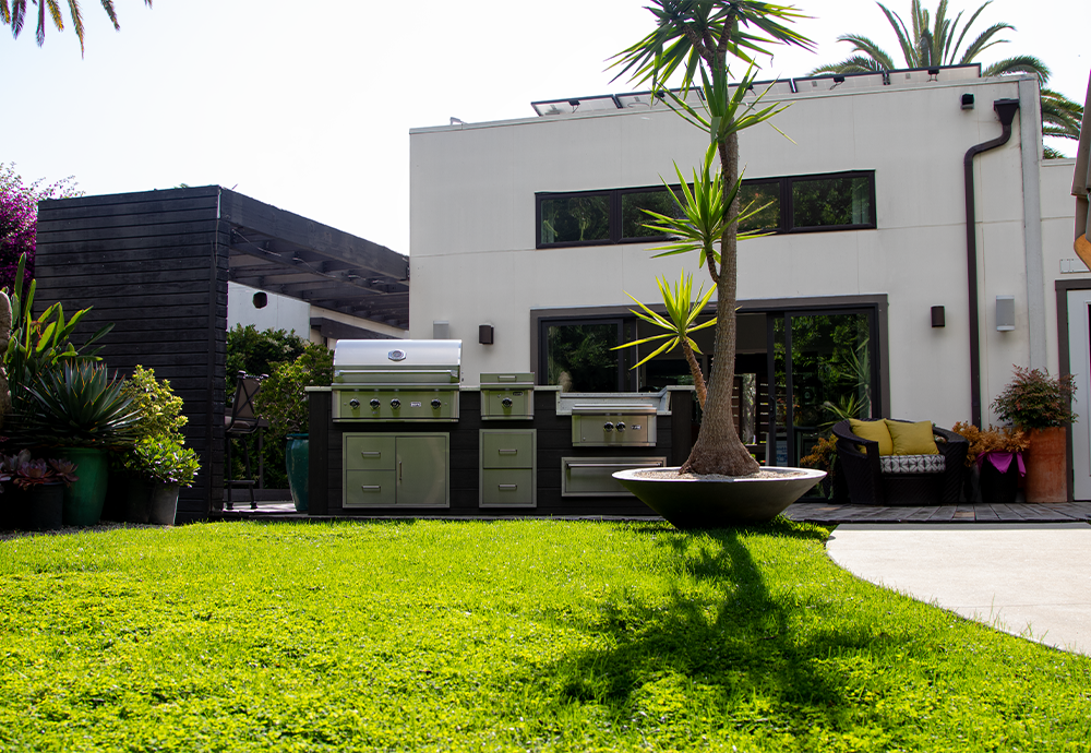 A modern backyard with a white house, black and green outdoor kitchen, palm trees, potted plants, a grassy lawn, and outdoor seating.