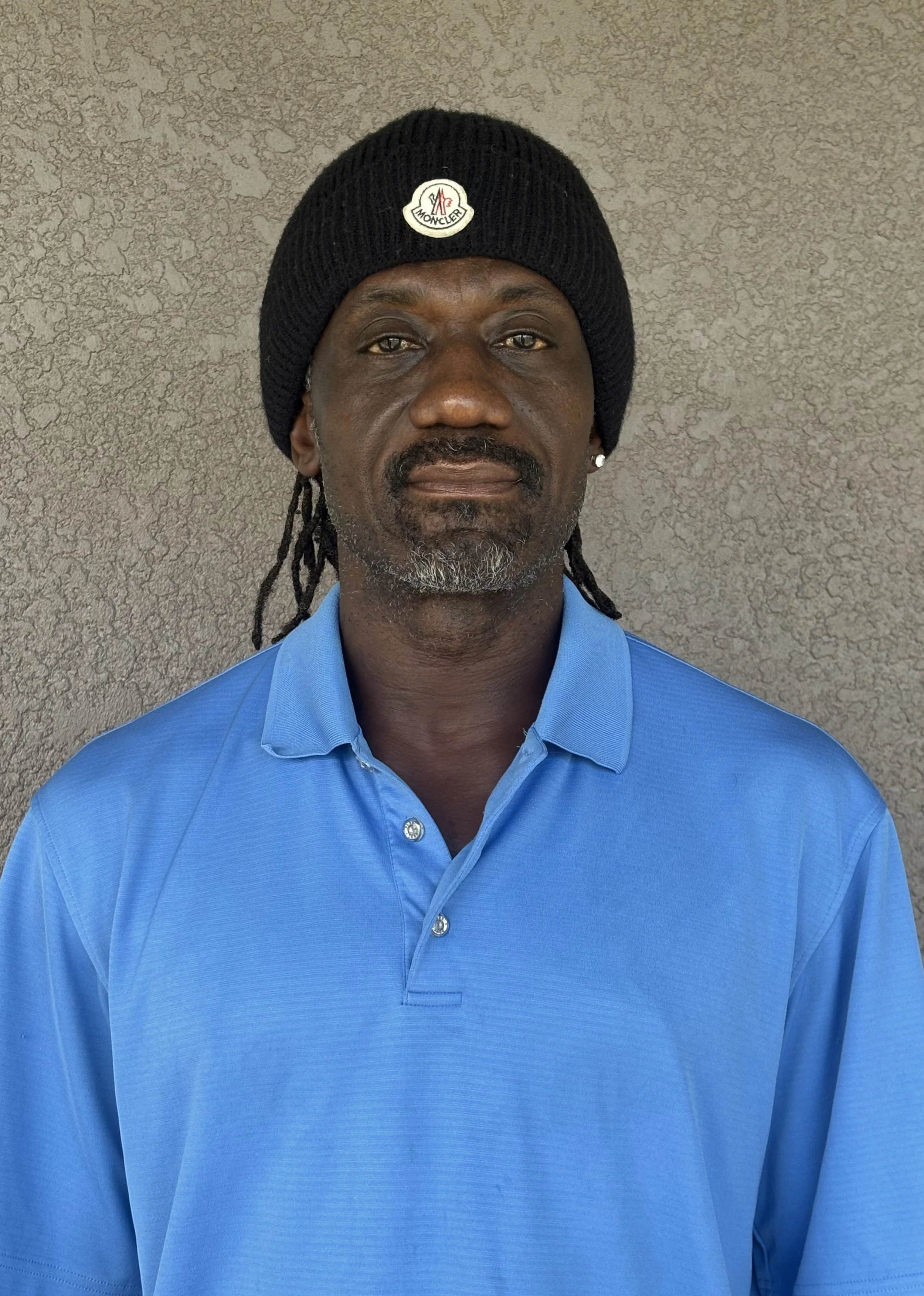 Man with dark skin, wearing a black beanie with a logo, a light blue polo shirt, and earrings, standing against a textured beige wall.